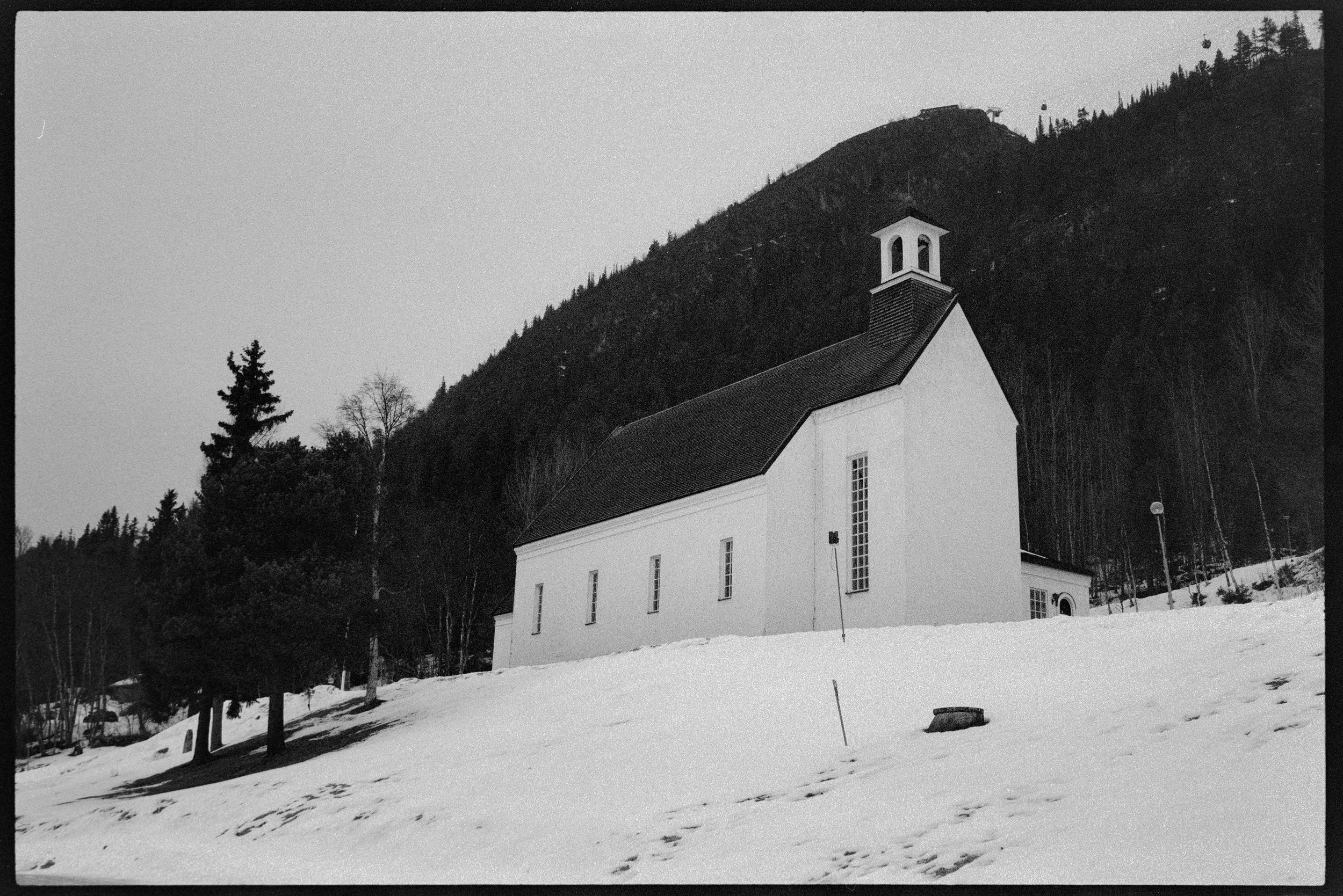 En vit kyrka med ett torn, omgiven av snötäckt mark och träd, med berg i bakgrunden.