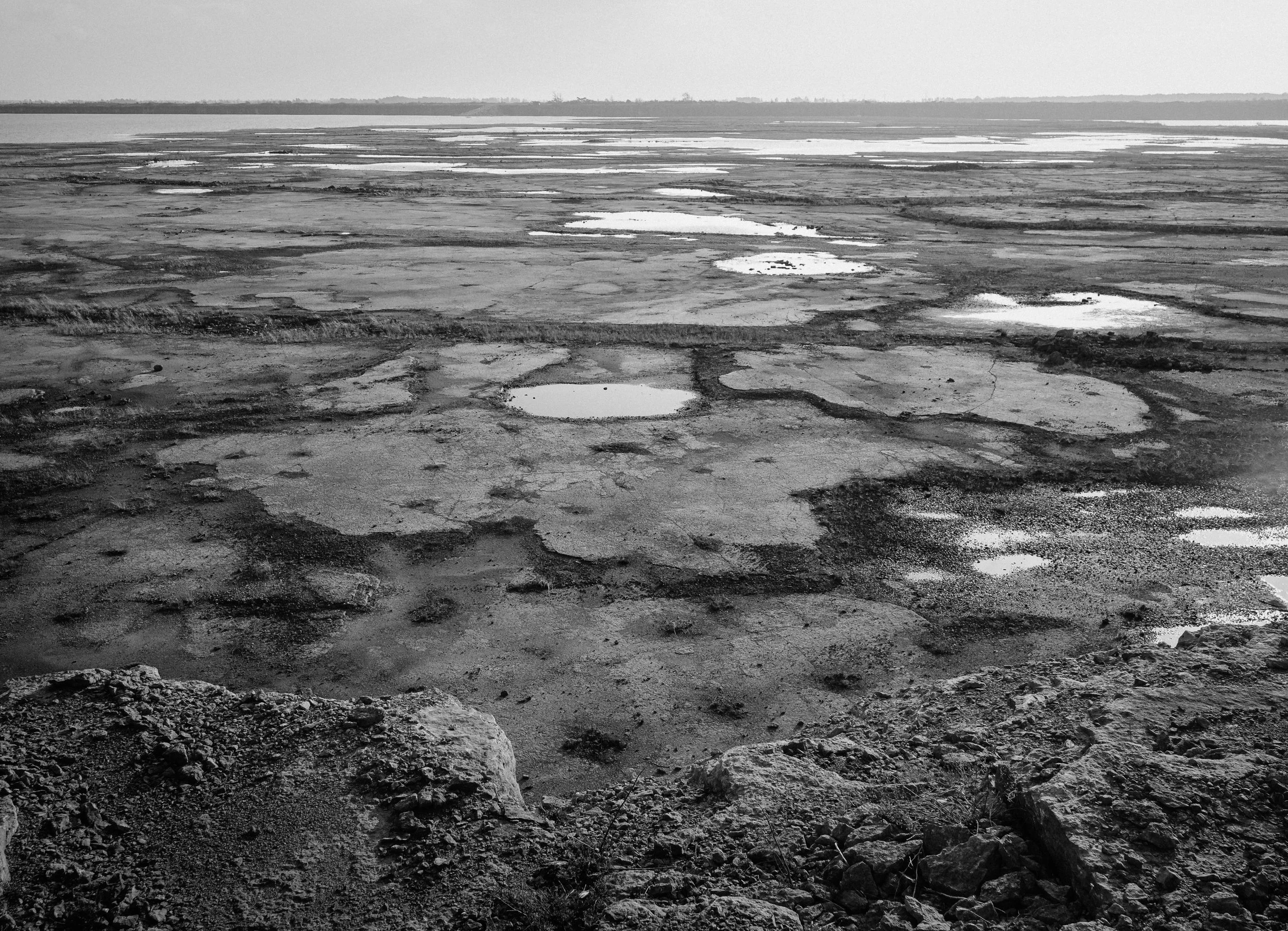 En utsikt över ett torrt område med små vattenpölar, grus och stenrös i förgrunden, och en horisontlinje i bakgrunden.