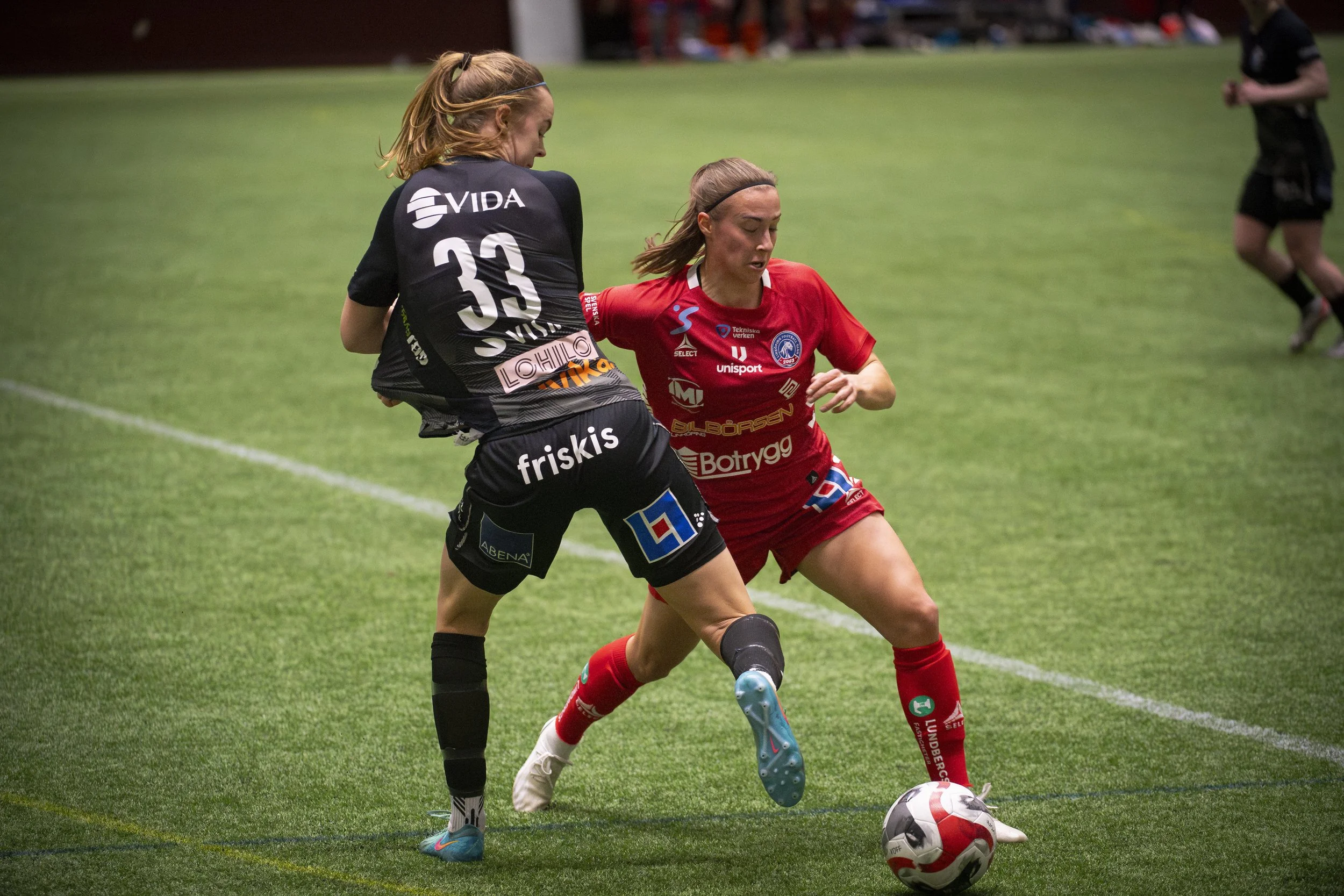 Två kvinnliga fotbollsspelare tävlar om bollen på planen. Den ena är i röd tröja och den andra i svart. De är nära varandra, med fokus på bollen.