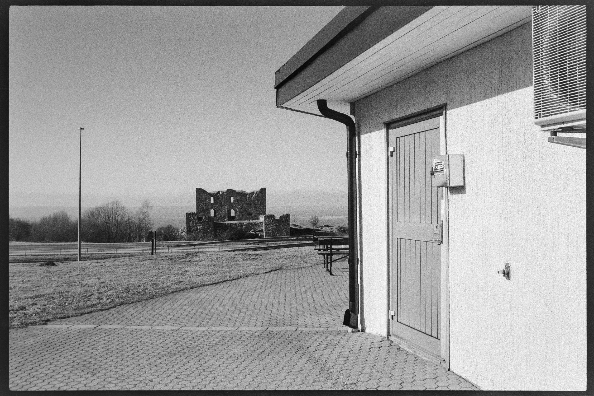 Svartvit bild av en byggnad med en dörr och ett rör utomhus, med en landsbygd i bakgrunden och ruinstillhörande struktur.