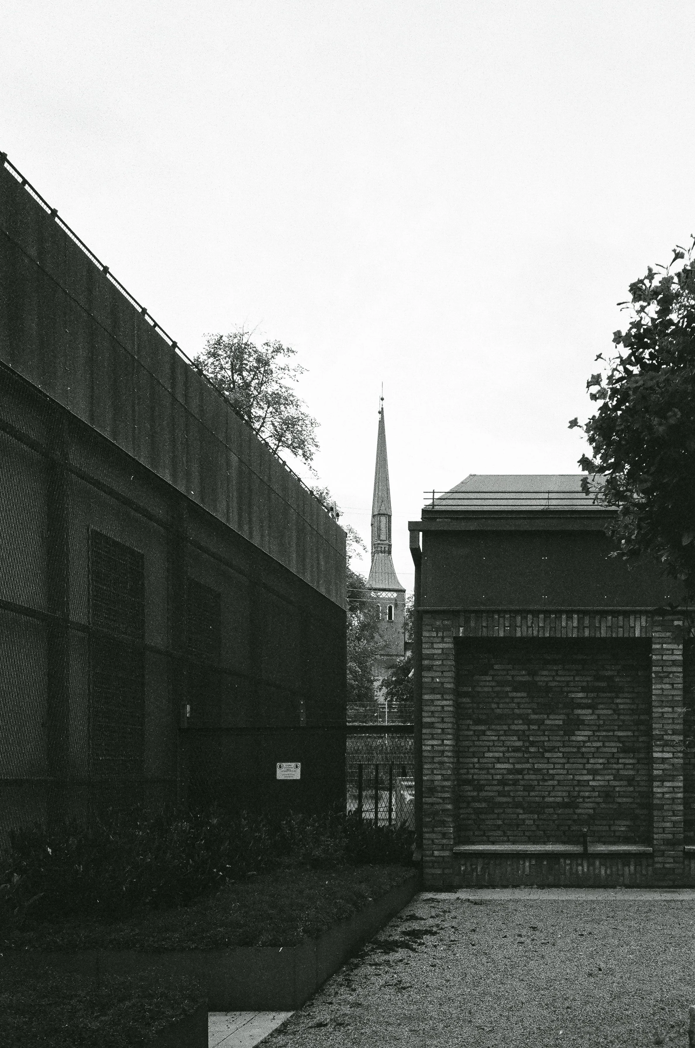 Svartvit bild av ett kyrktorn som sticker upp mellan två byggnader, med en tydlig, spetsig takremsa och en liten balkong nertill.