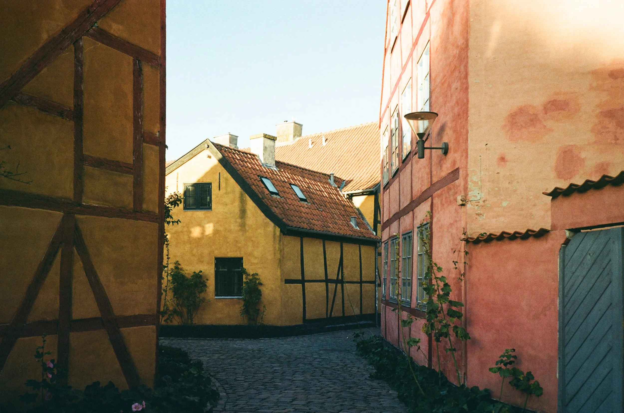 En smal kullerstensgränd mellan färgglada gamla hus, ett gult hus till vänster och ett rosa till höger, med en solig himmel i bakgrunden.