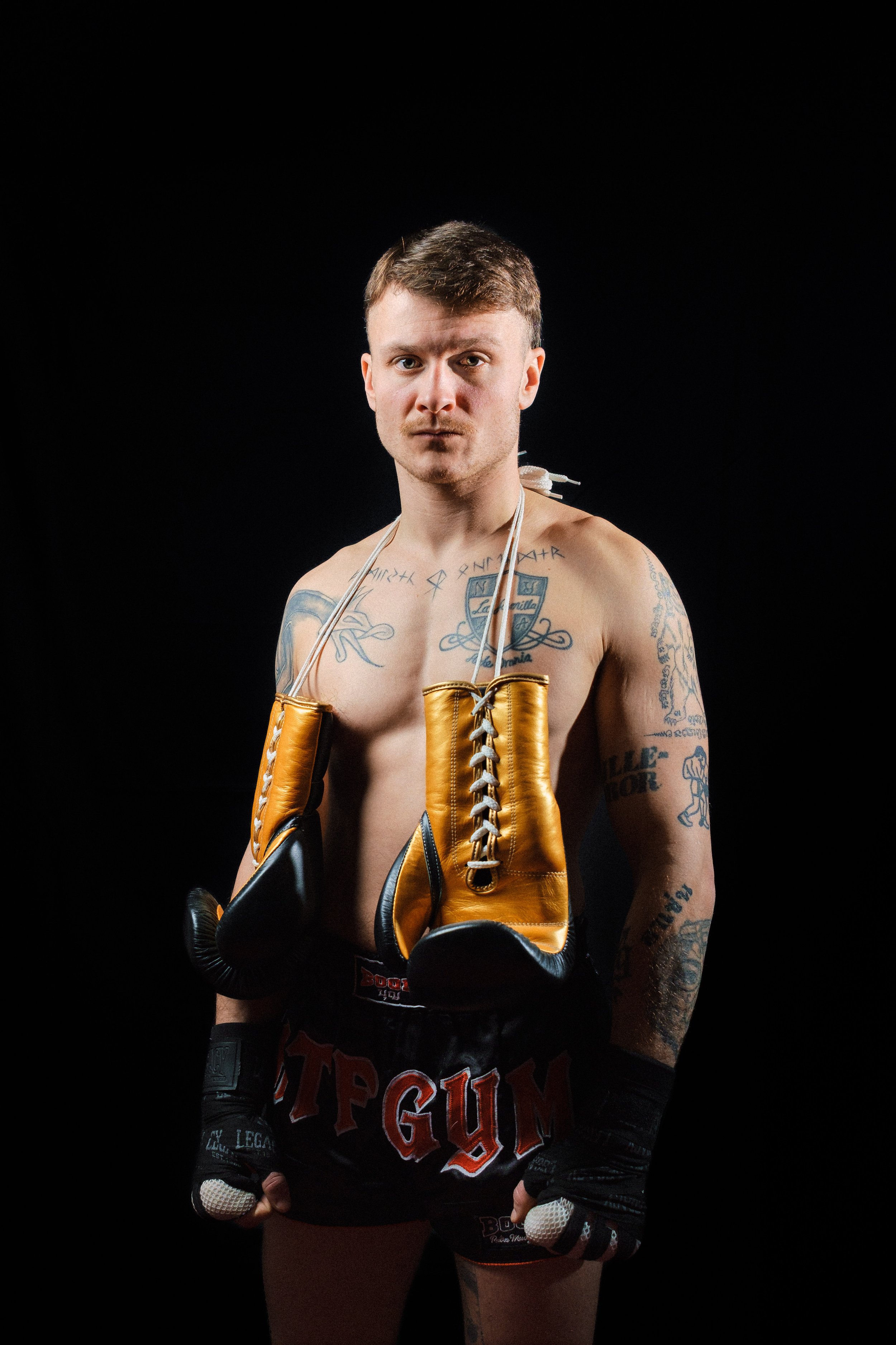 En man i MMA-kläder med boxhandskar och ett par gummistövlar hänger runt halsen, står mot svart bakgrund.