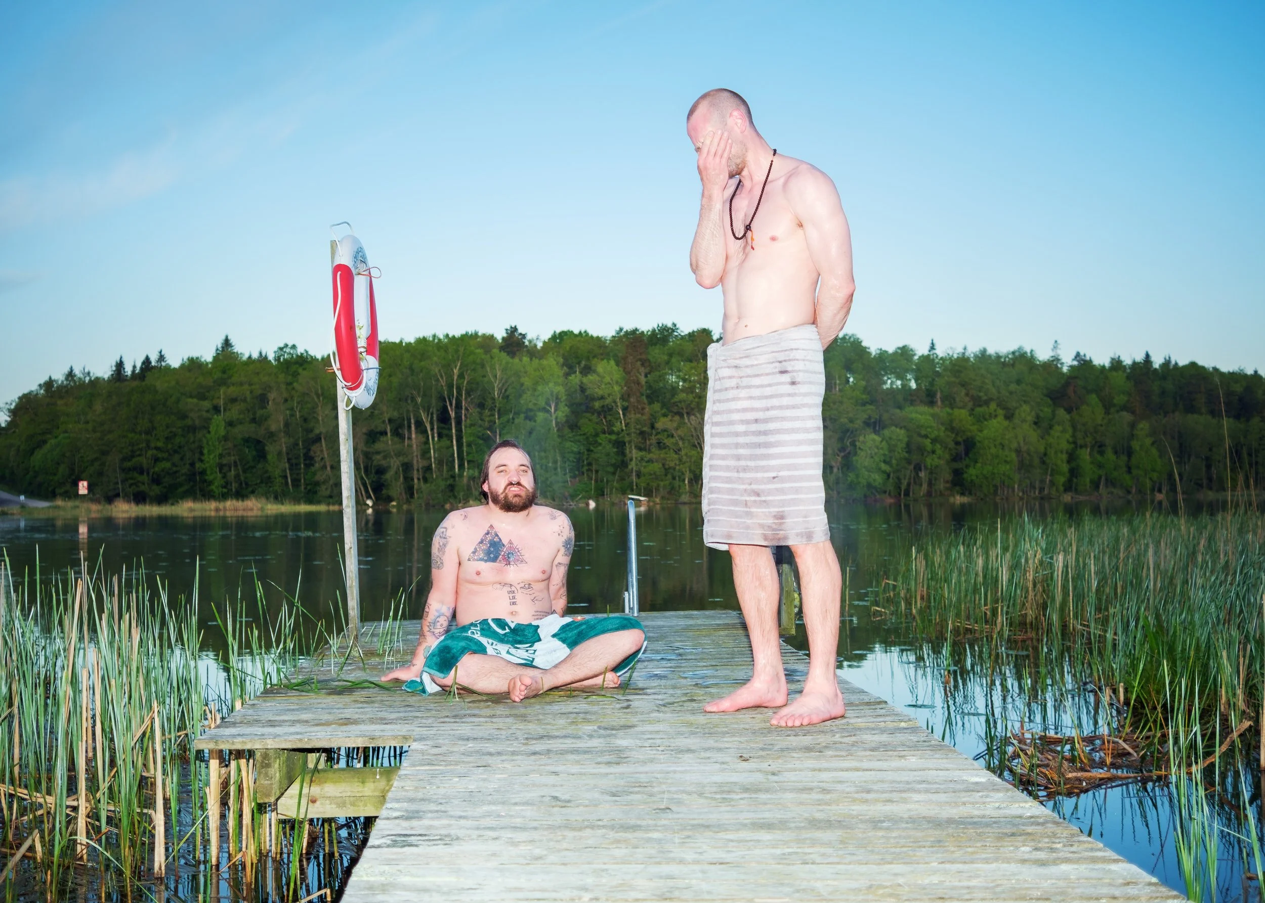 Två män på en flytbrygga vid en sjö, en av dem sitter med korslagda ben och den andra står med handen över ansiktet.