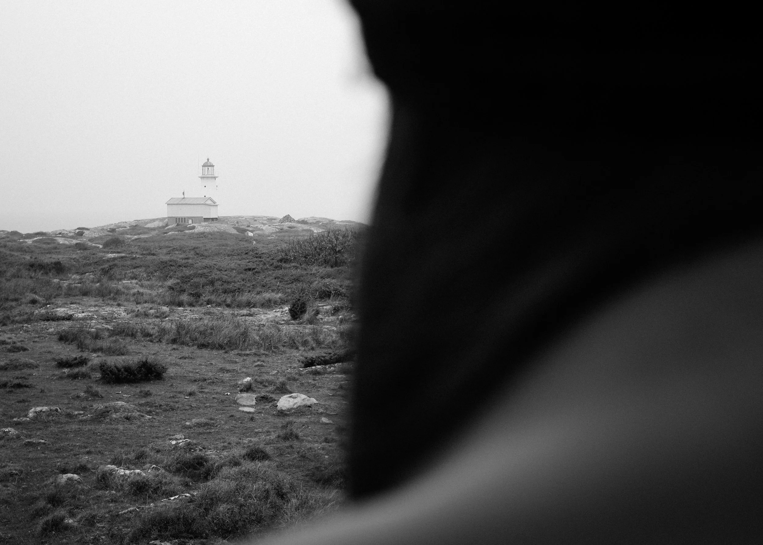 En svartvit bild av ett landskap med ett hus och en fyr i horisonten, delvis täckt av en mörk skugga i förgrunden.