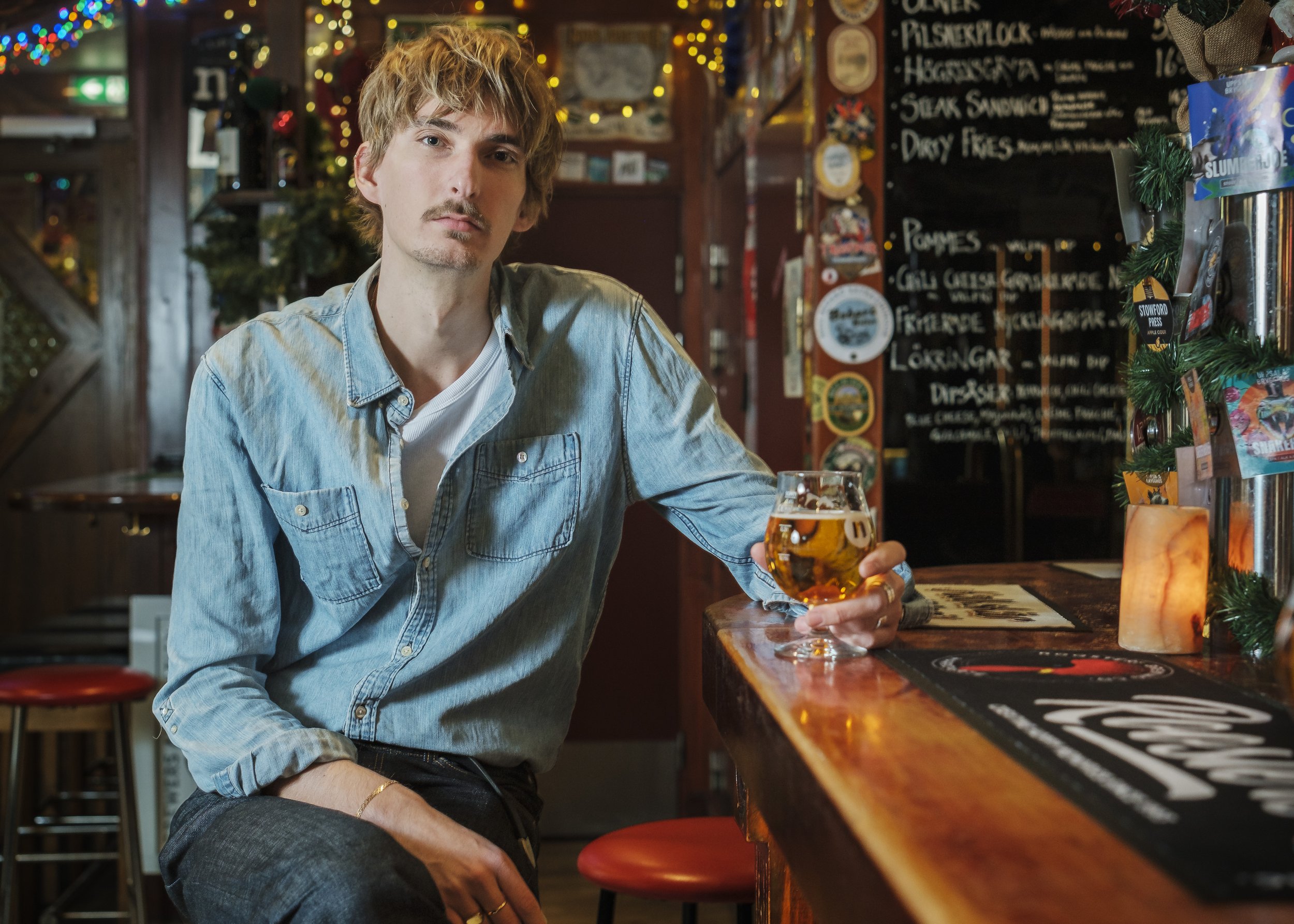 En man som sitter vid baren i en pub, håller i ett glas öl, och tittar mot kameran. Bakom honom finns en menybricka och juldekorationer.
