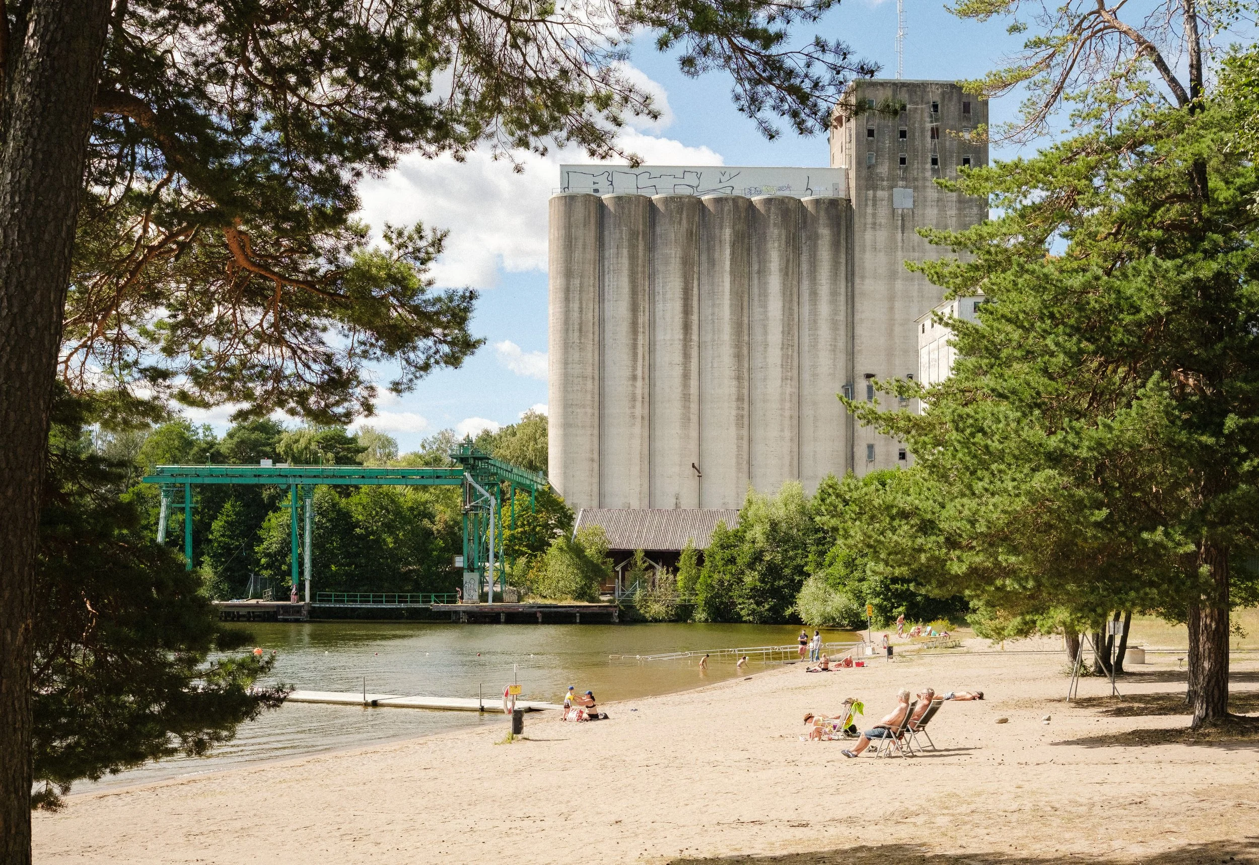 Sandstrand vid en sjö med personer som solar och badar, omgiven av gröna träd och en stor industriell byggnad i bakgrunden.