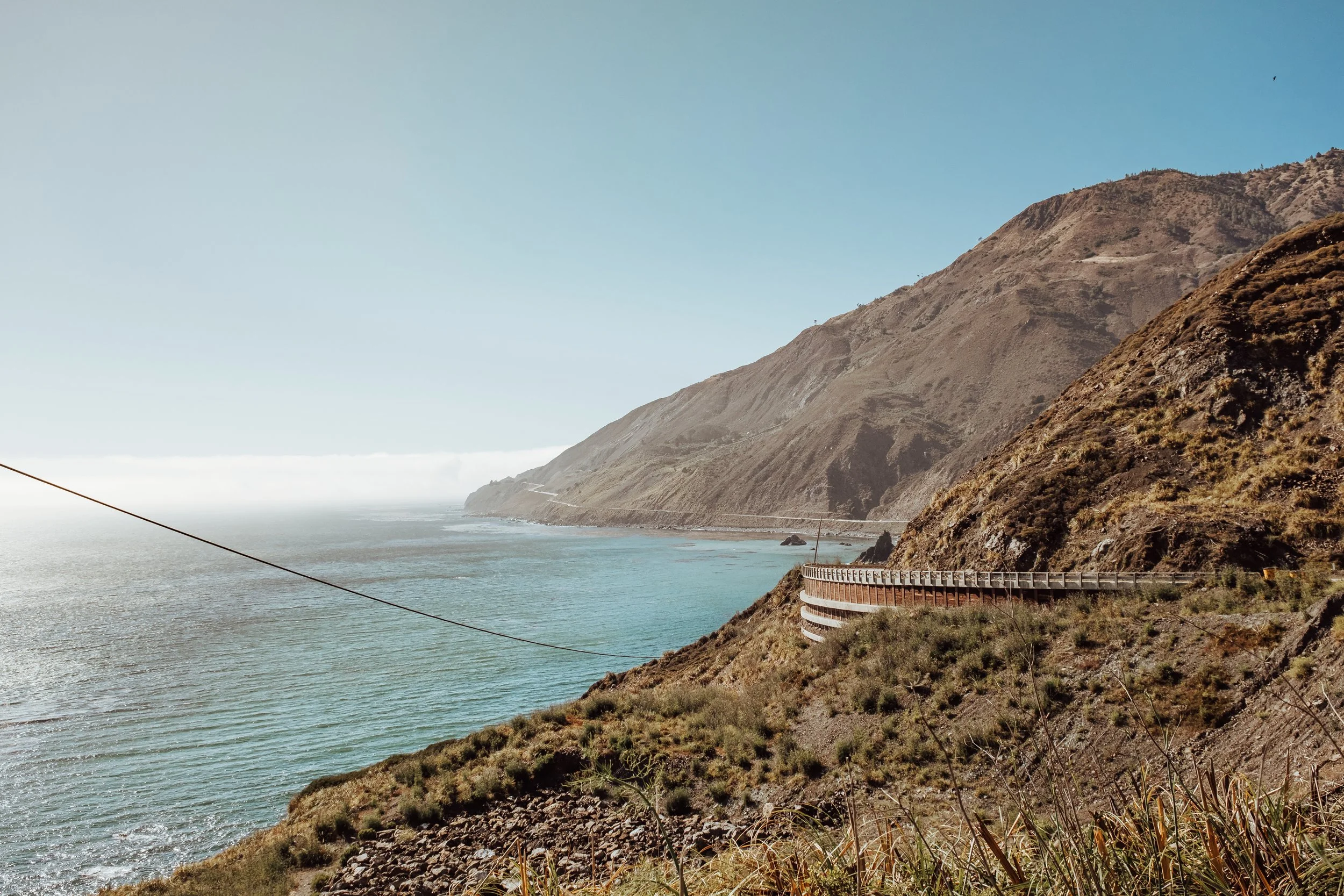 En kust med branta klippor och en bilväg som slingrar sig längs berget, med havet och en klar blå himmel i bakgrunden.