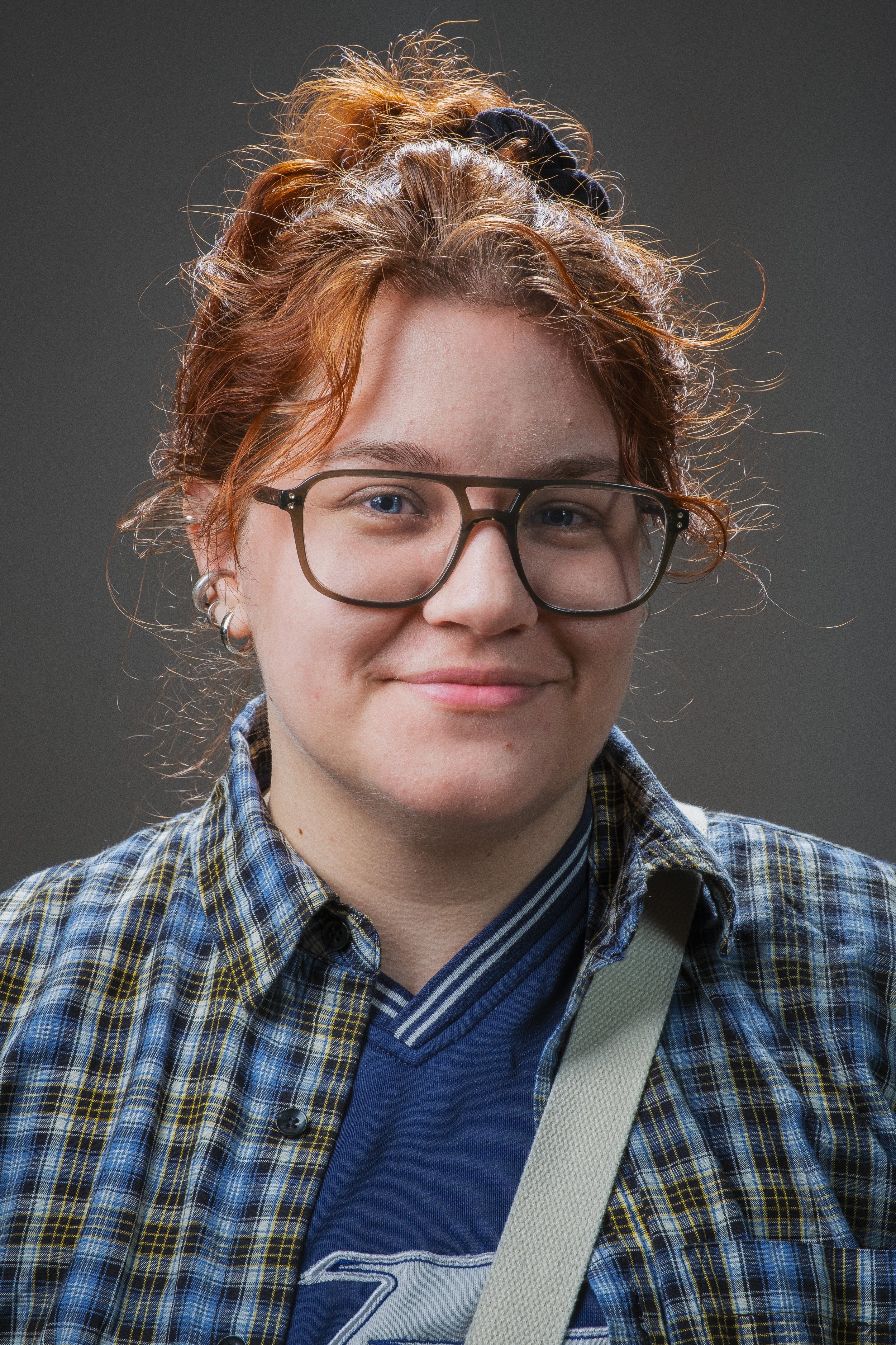Porträtt av en ung person med rödaktigt hår, glasögon, prickig skjorta och blå t-shirt, mot en grå bakgrund.