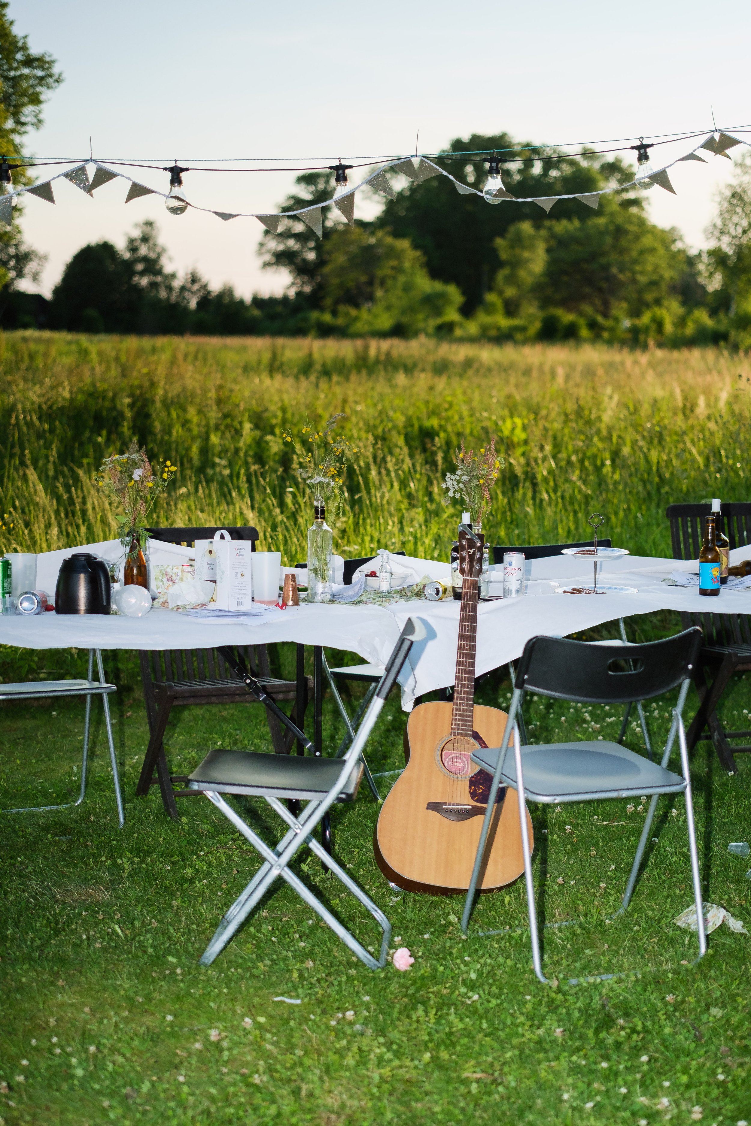 Utemiddag eller festbord täckt med en vit duk, dekorerat med vaser och flaskor med blommor, omgiven av stolar, en gitarr står på marken, i bakgrunden ett grönt fält och träd, hängande ljusslinga över bordet.