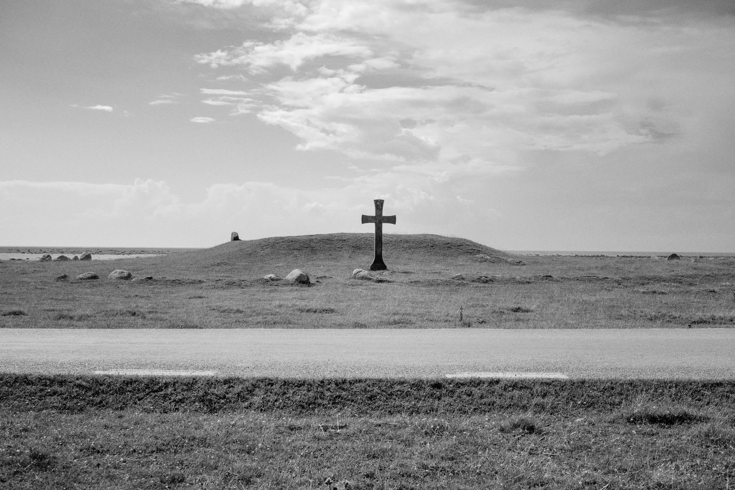 En svart kors på en kulle omgiven av gräs, med en väg i förgrunden och moln på himlen.