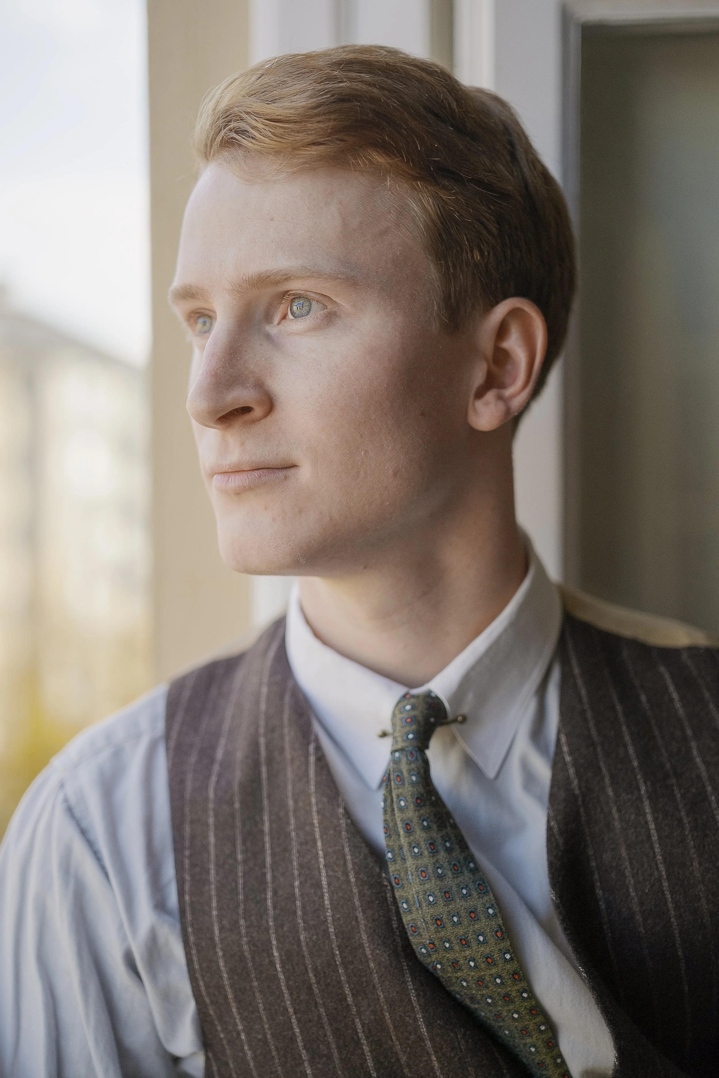 En man med ljust skin och ljusblå ögon tittar ut genom ett fönster, klädd i vit skjorta, mörk väst och en mönstrad slarv. Han har rödbrunt hår och ett lugnt uttryck.