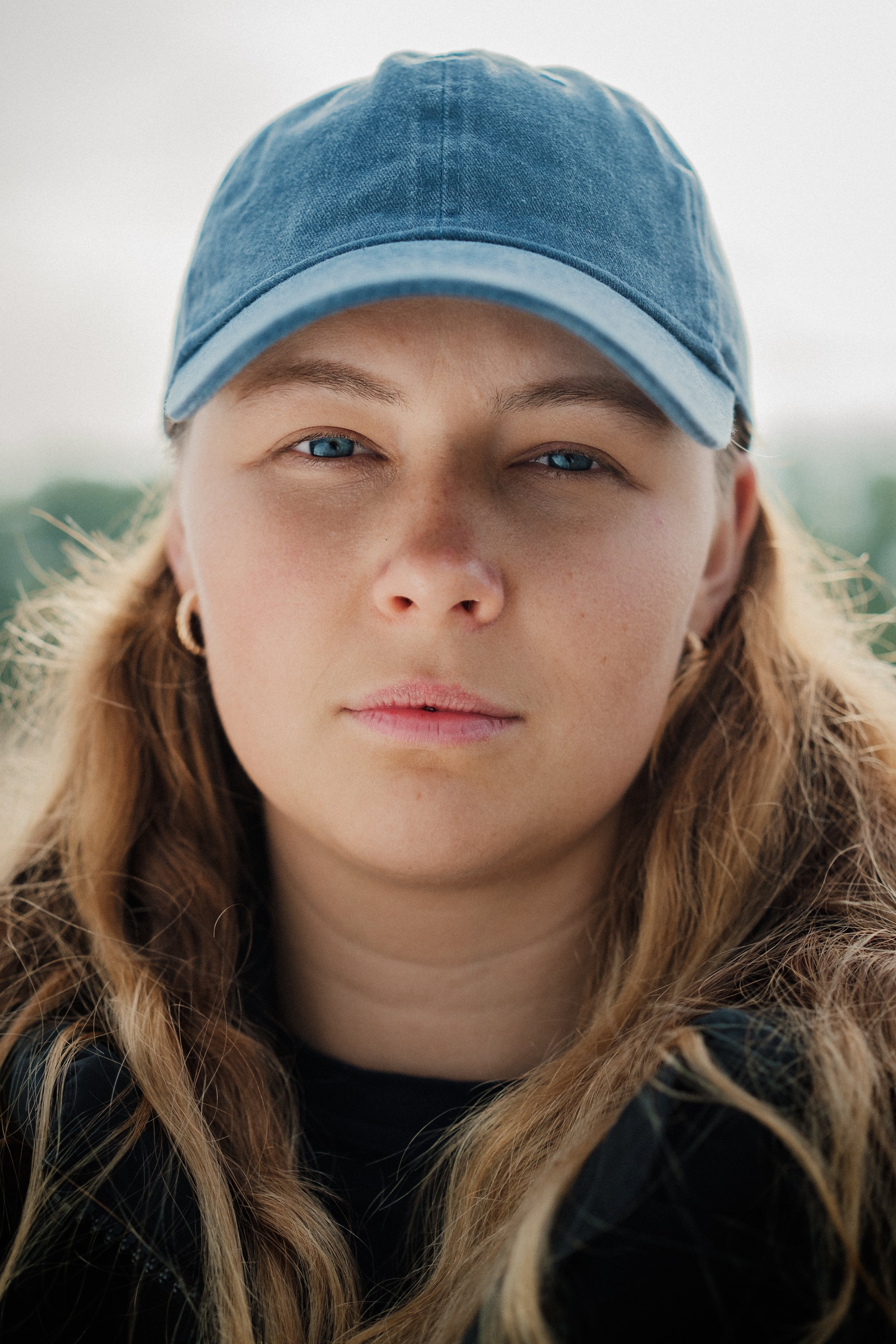 Närbild av en ung kvinna med blå ögon, långt lockigt hår och mörk tröja, som bär en blå keps.