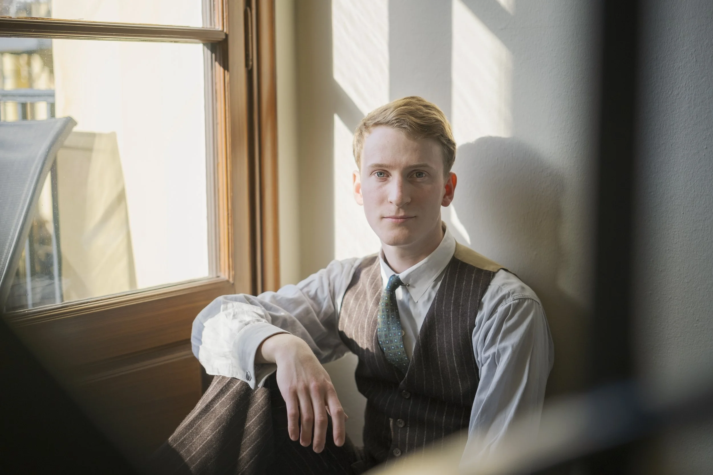 En ung man i vit skjorta, mörkbrun väst och slips sitter vid ett fönster och tittar mot kameran.