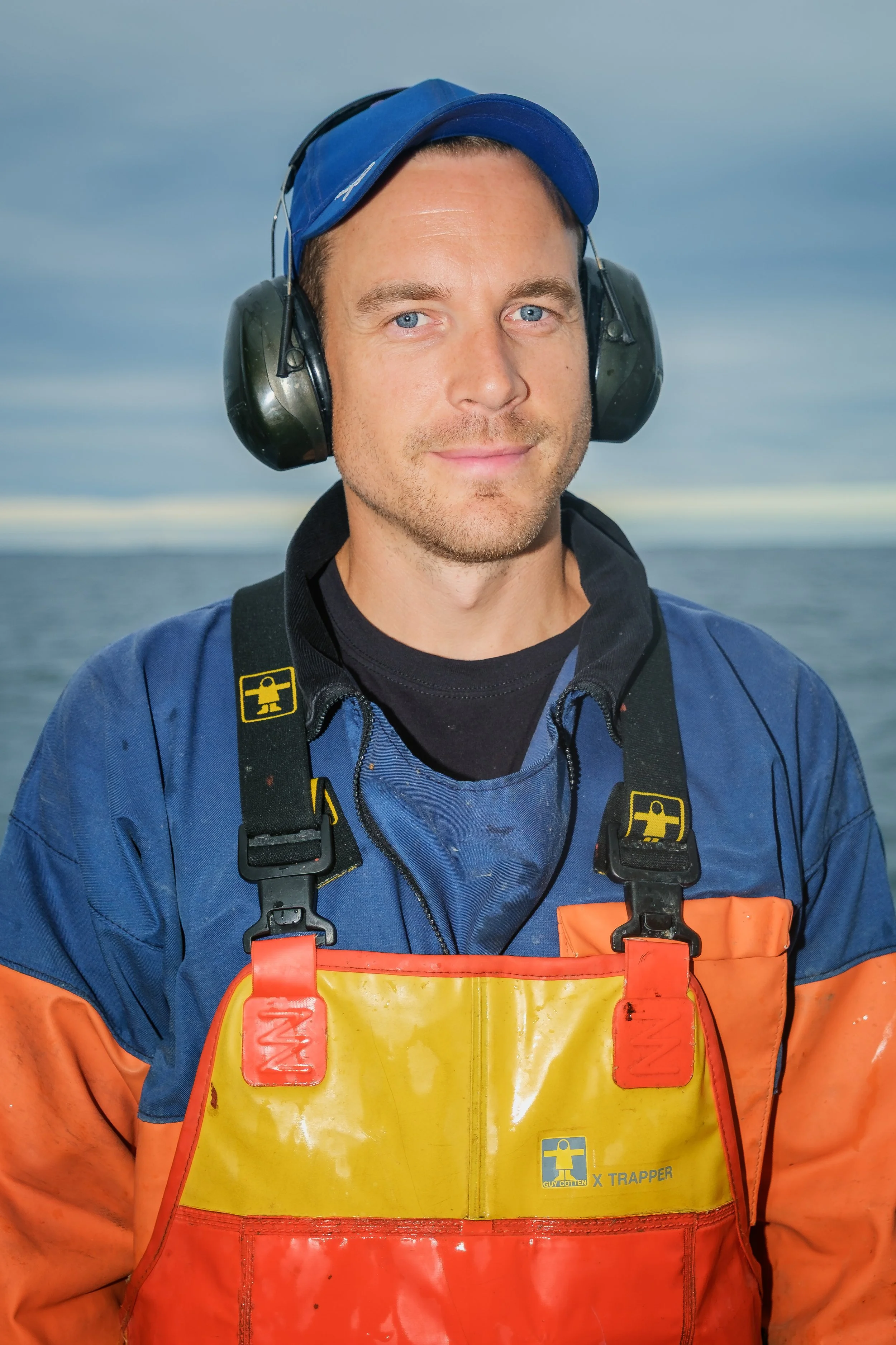 En man i kustnära arbetskläder, med hörselskydd, blå keps och en orange och gul väst, står vid havet och ler.