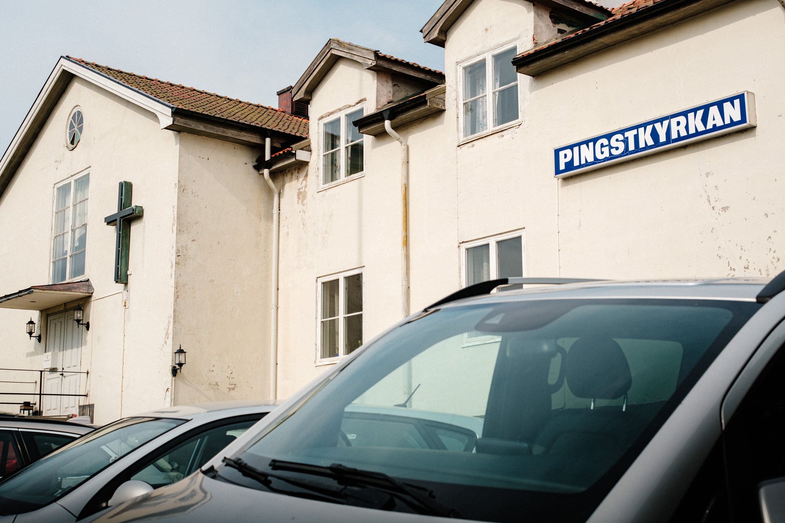 En vit kyrkobyggnad med ett blått skylt som säger Pingstkyrkan, parkering med bilar framför kyrkan.