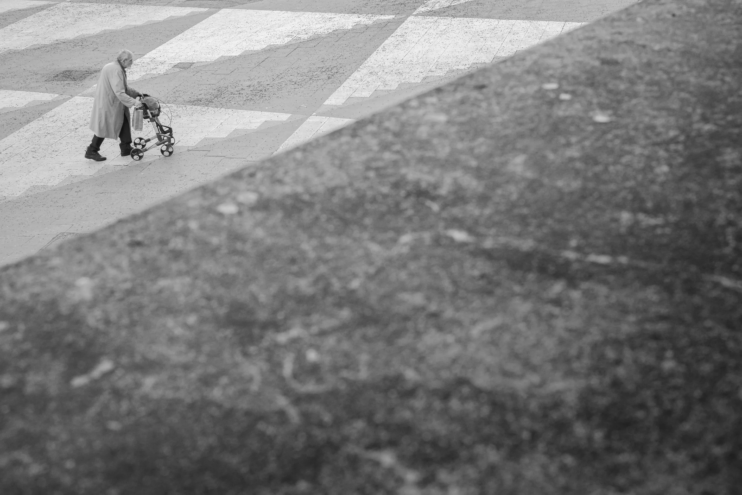 En äldre person går med en rullator på en trottoar.