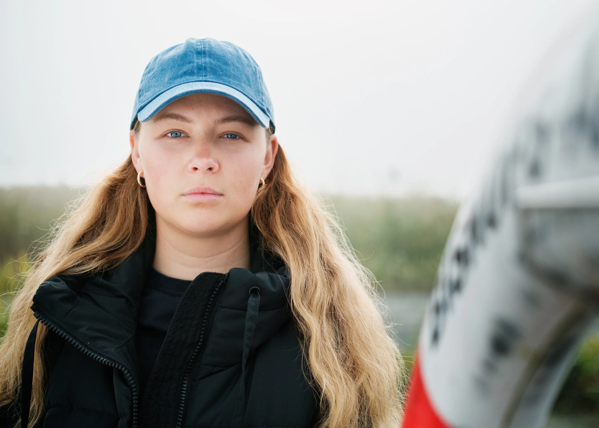 En ung kvinna med lång, lockigt blond hår, blå ögon, iklädd svart jacka och blå keps.