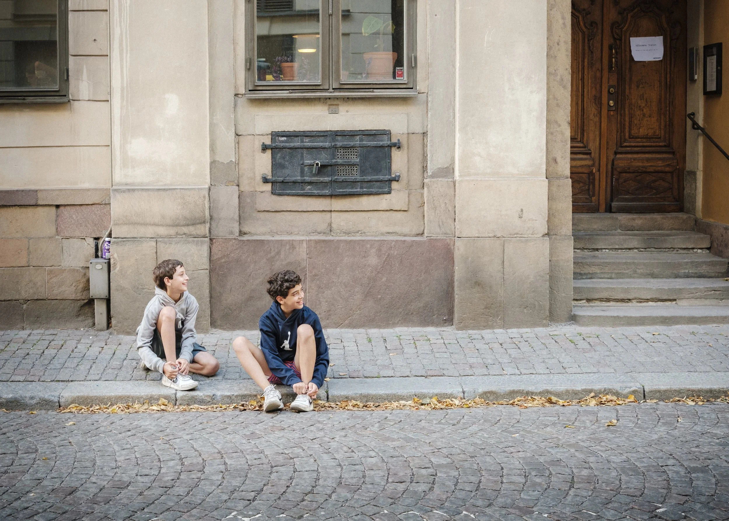 Två pojkar sitter på trottoaren utanför en gammal byggnad med stenfasad och träport. De är avslappnade och ser ut att ha en pratstund.