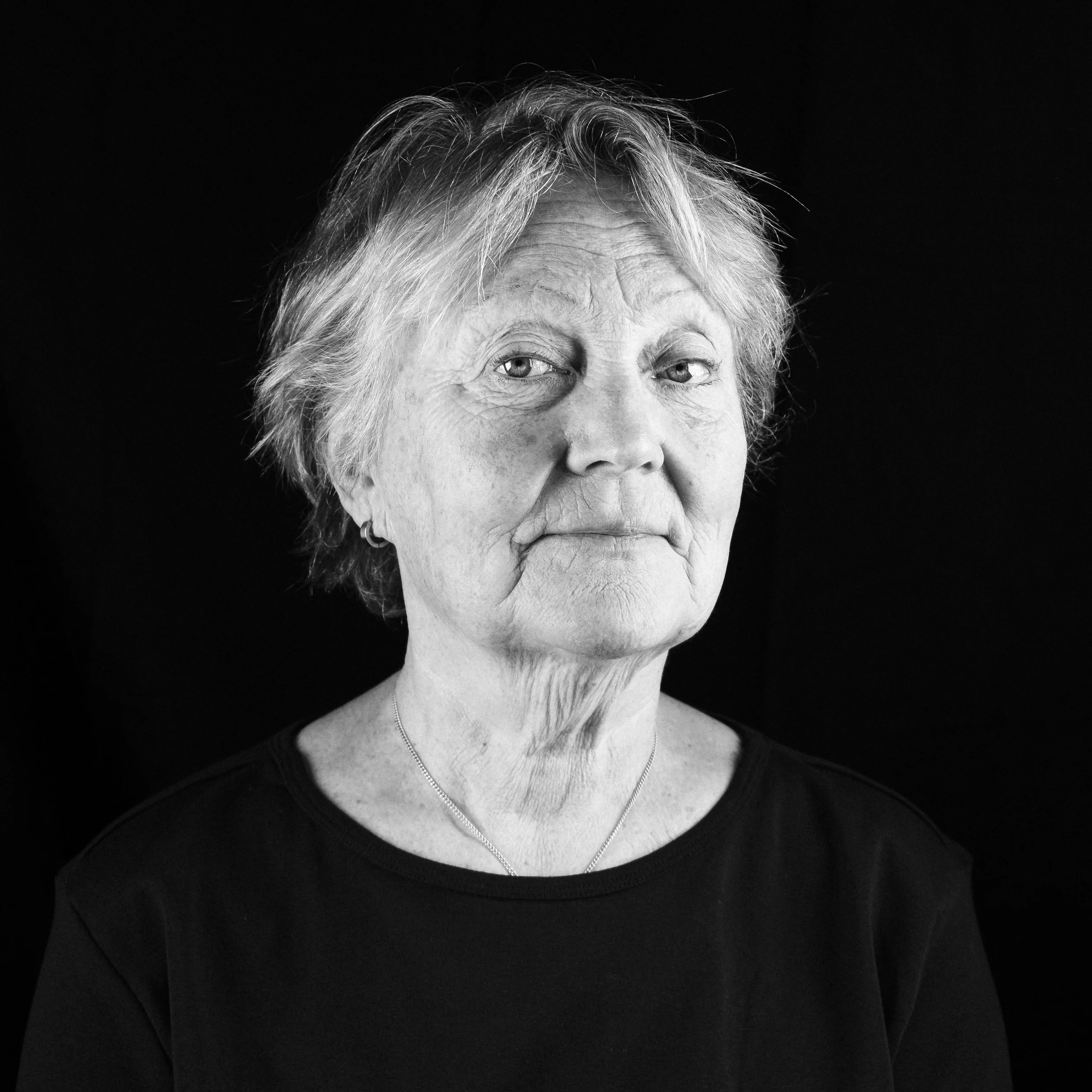Svartvitt porträtt av en äldre kvinna med kort, vågigt hår och earring, klädd i svart tröja, mot en mörk bakgrund.