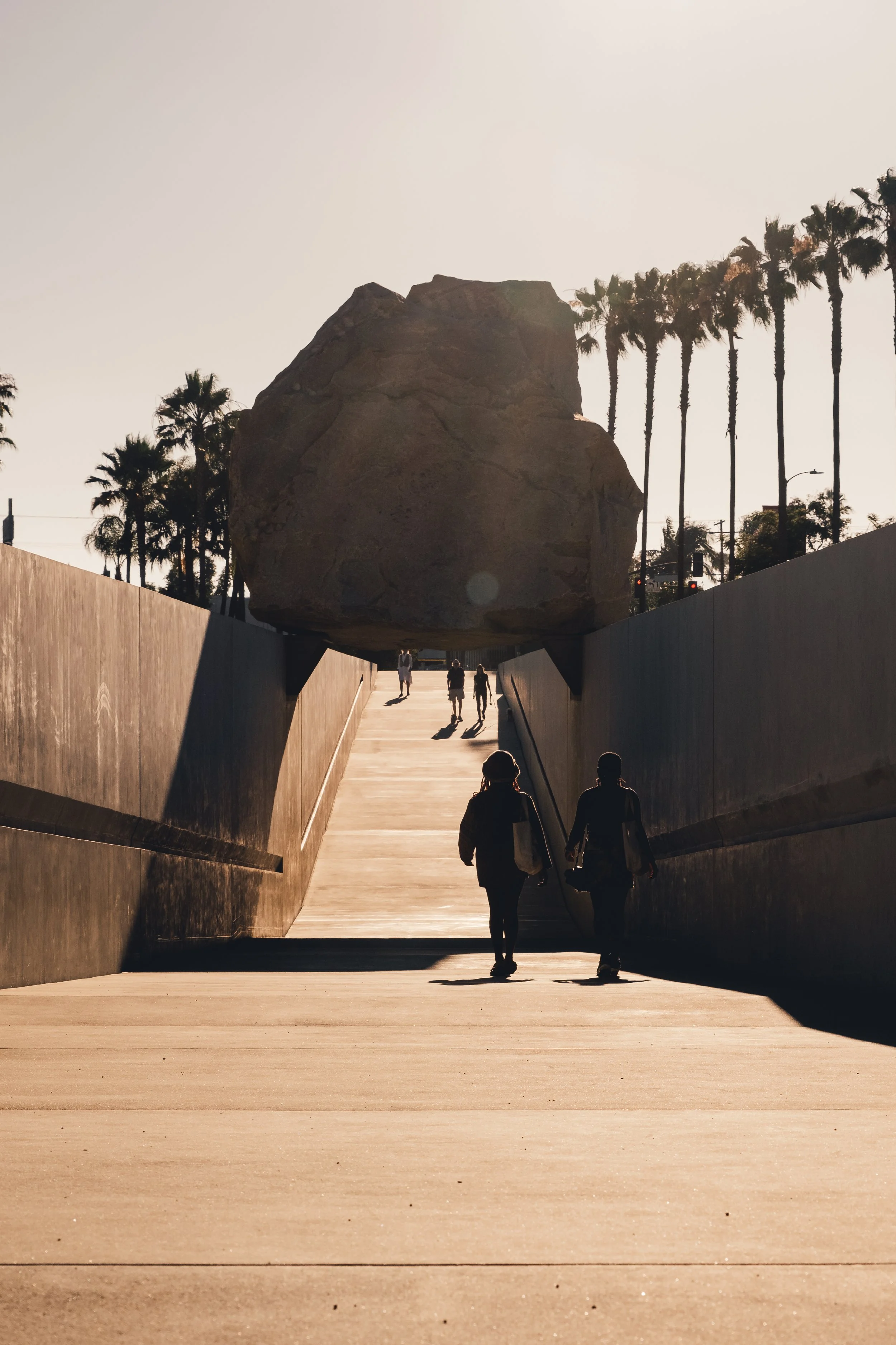 Två personer går på en trappa med ett stort klipp ovanför dem, omgivna av palmer och en ljus himmel.