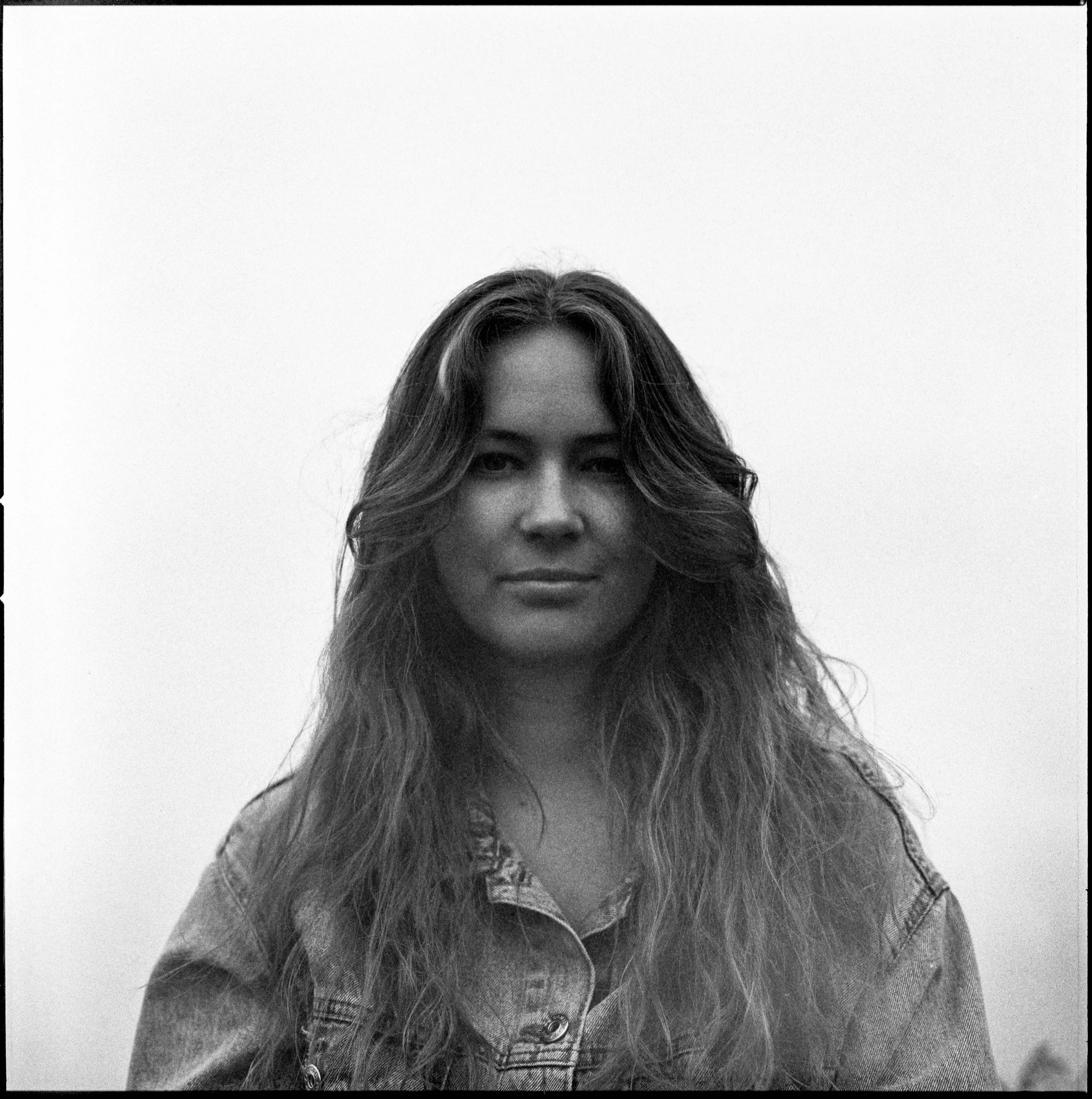 Svartvitt fotografi av en ung kvinna med långt, lockigt hår, iförd denimjacka, som tittar mot kameran. Bakgrunden är en enkel, tom vägg.