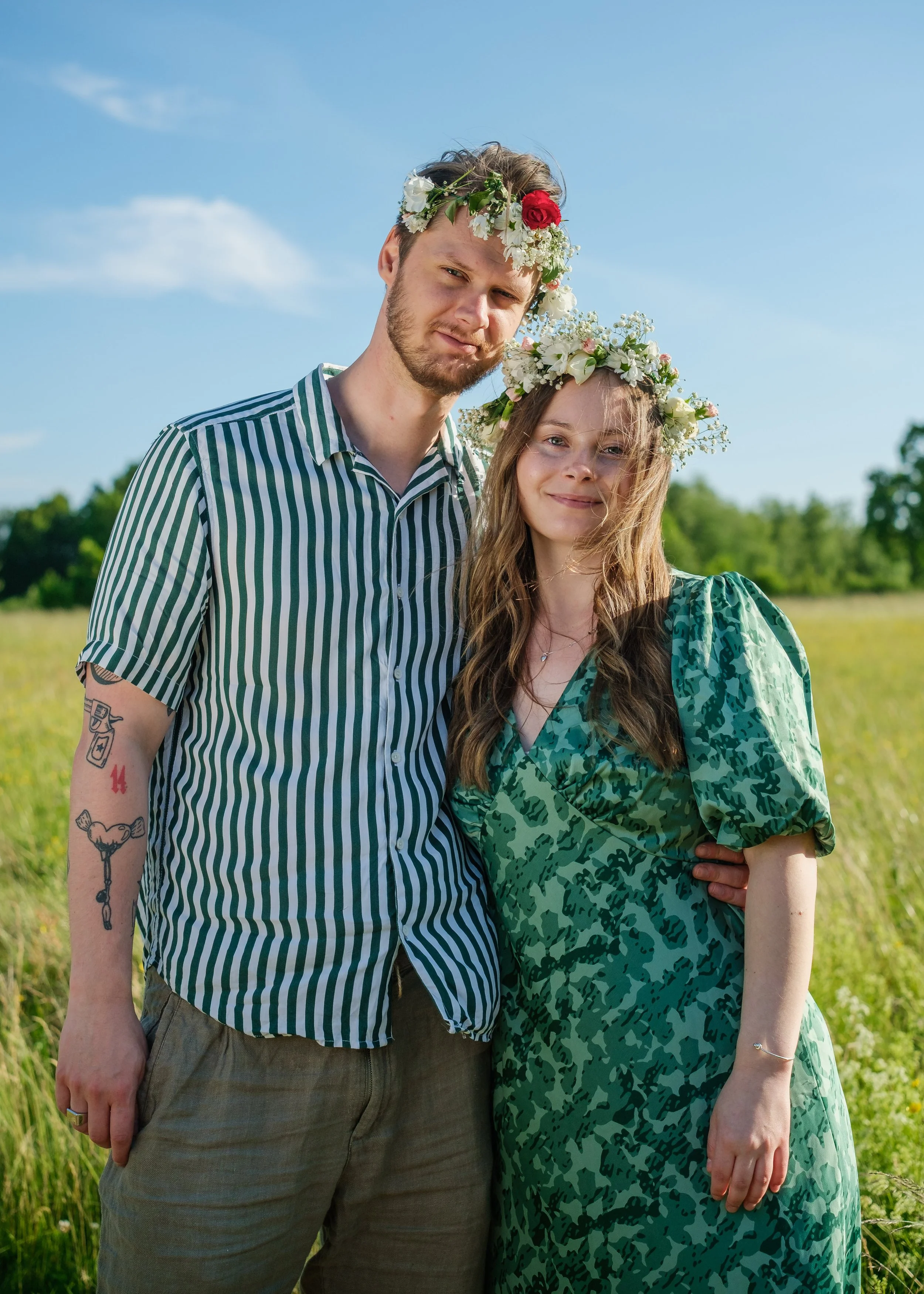 En man och en kvinna står i ett fält under en klarblå himmel, båda bär kransar av blommor på huvudet, insvepta i gröna kläder.