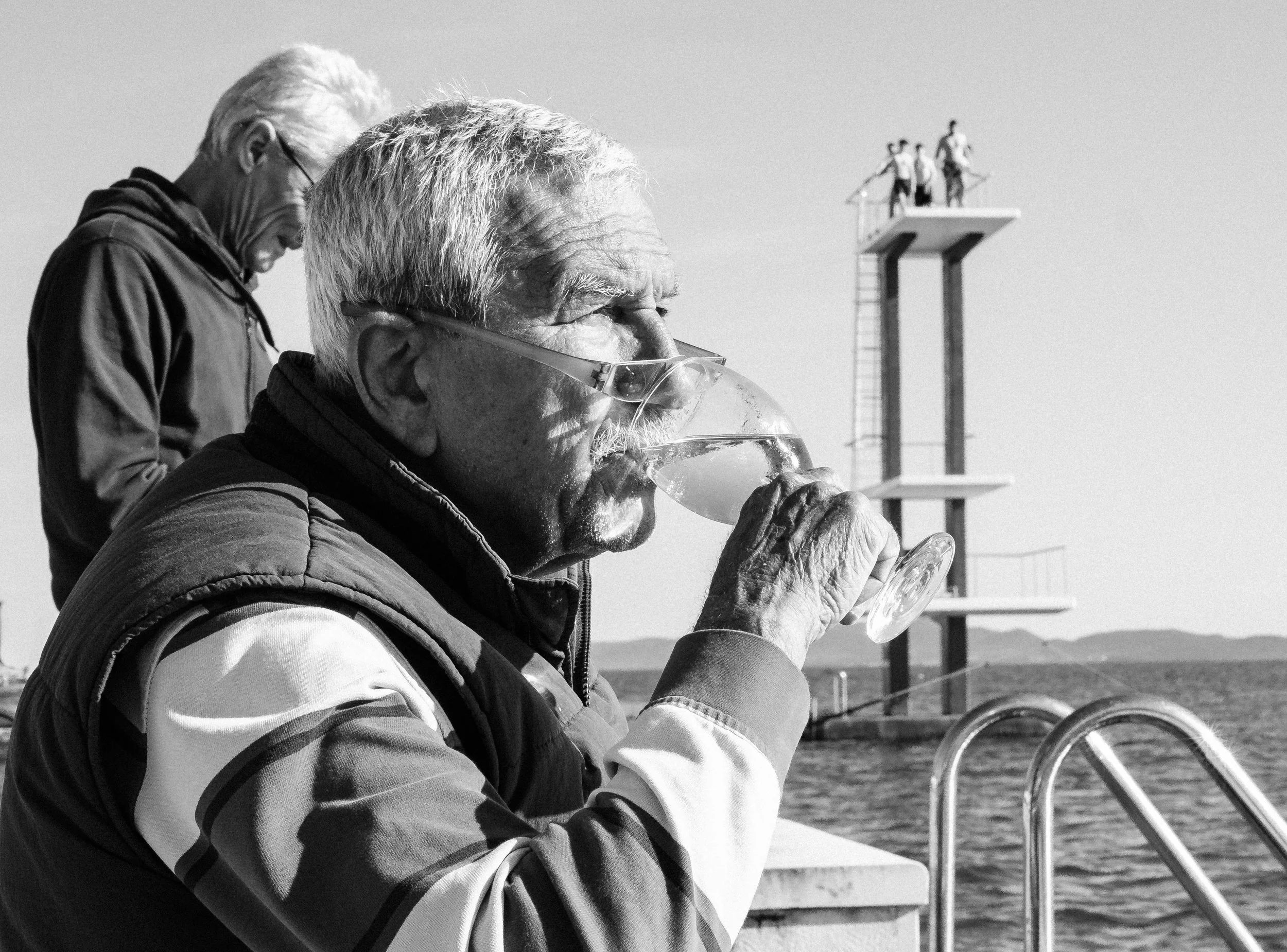 En äldre man dricker ett glas vin vid en pool, bakgrund med ett hopptorn och en man som tittar ner.