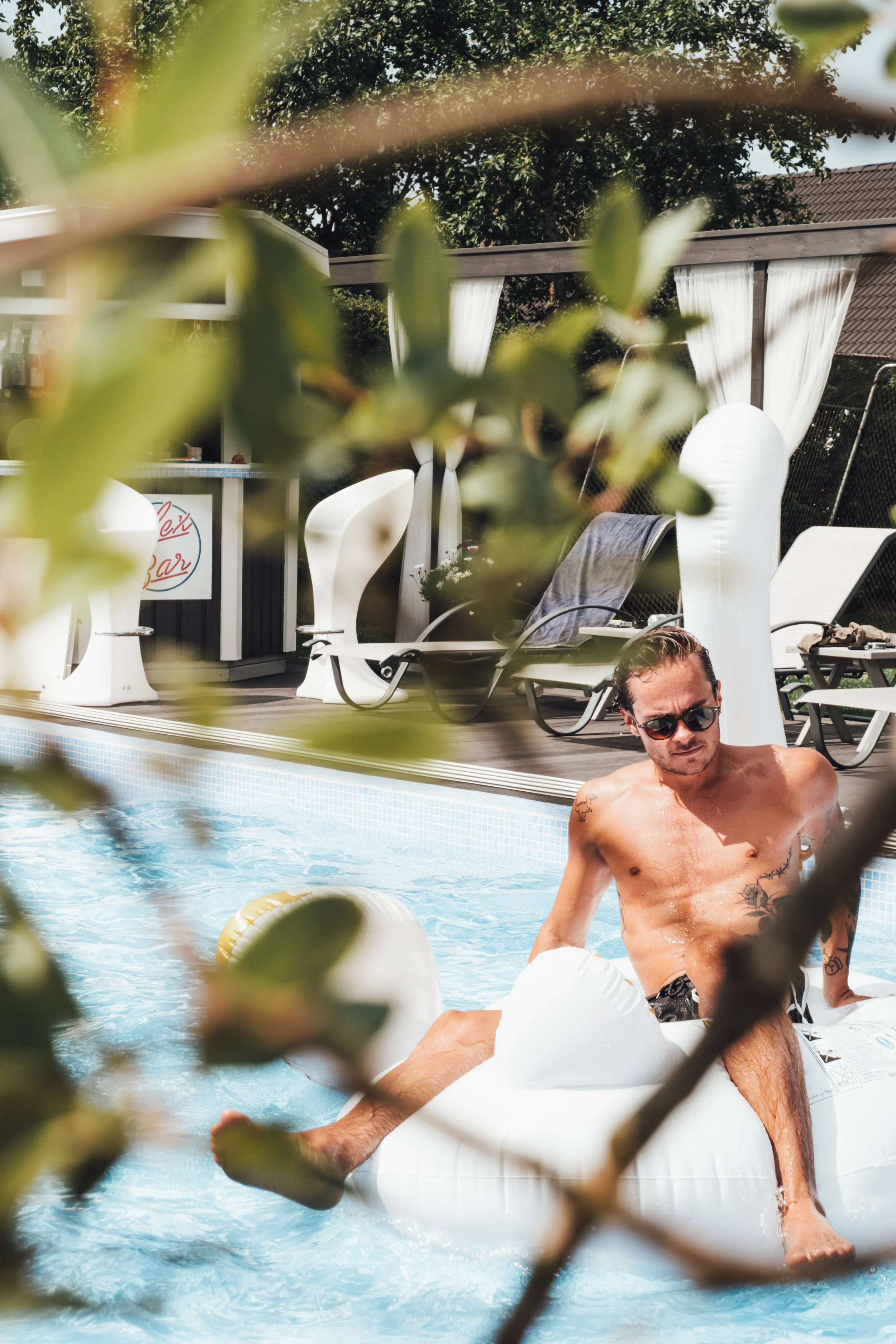 En man i solglasögon som solar sig på en uppblåsbar flamingo i en swimmingpool, omgiven av gröna växter och utomhusmöbler.