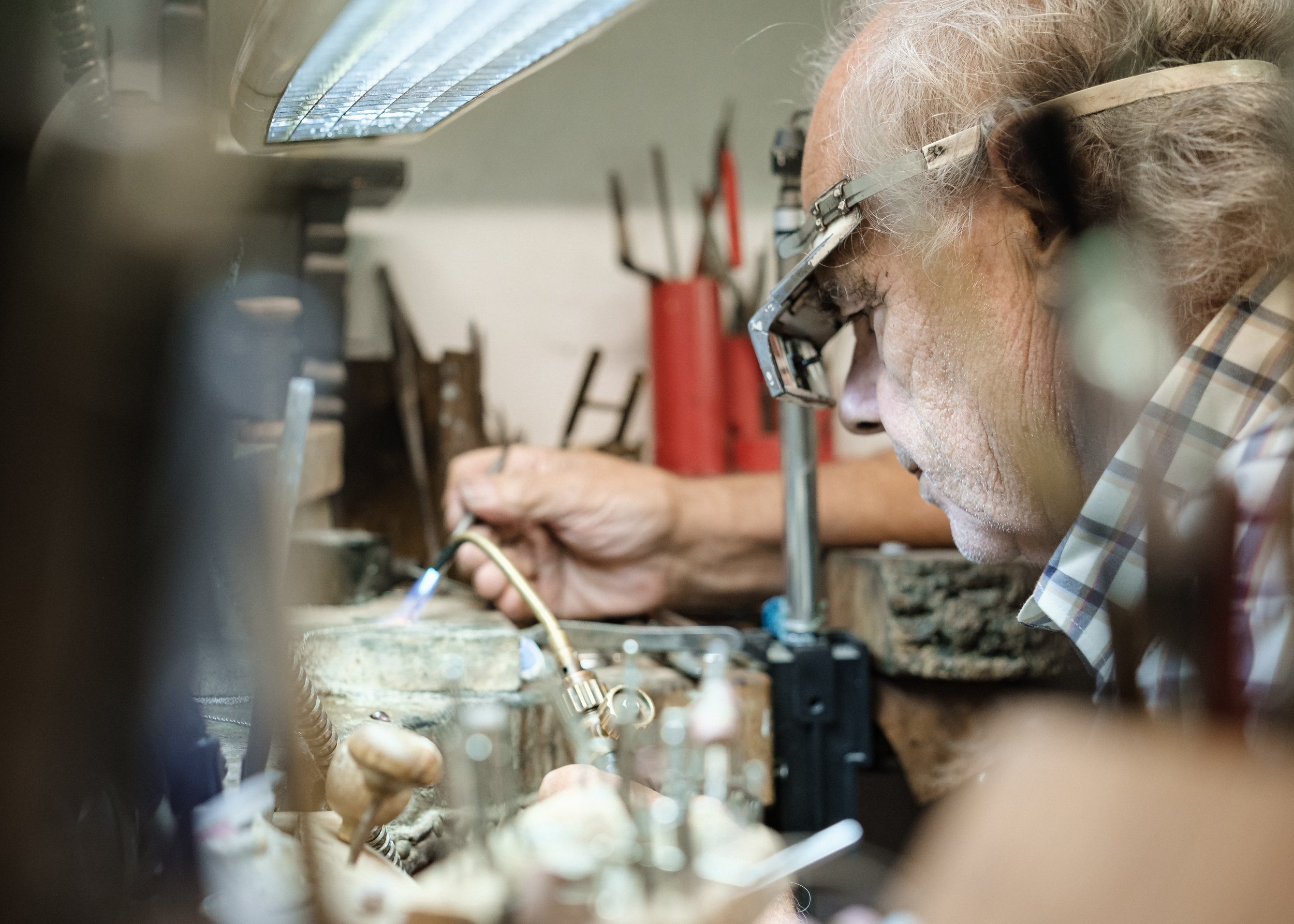 En äldre man med glasögon arbetar med en lödning på en arbetsbänk i ett verkstadsmiljö.