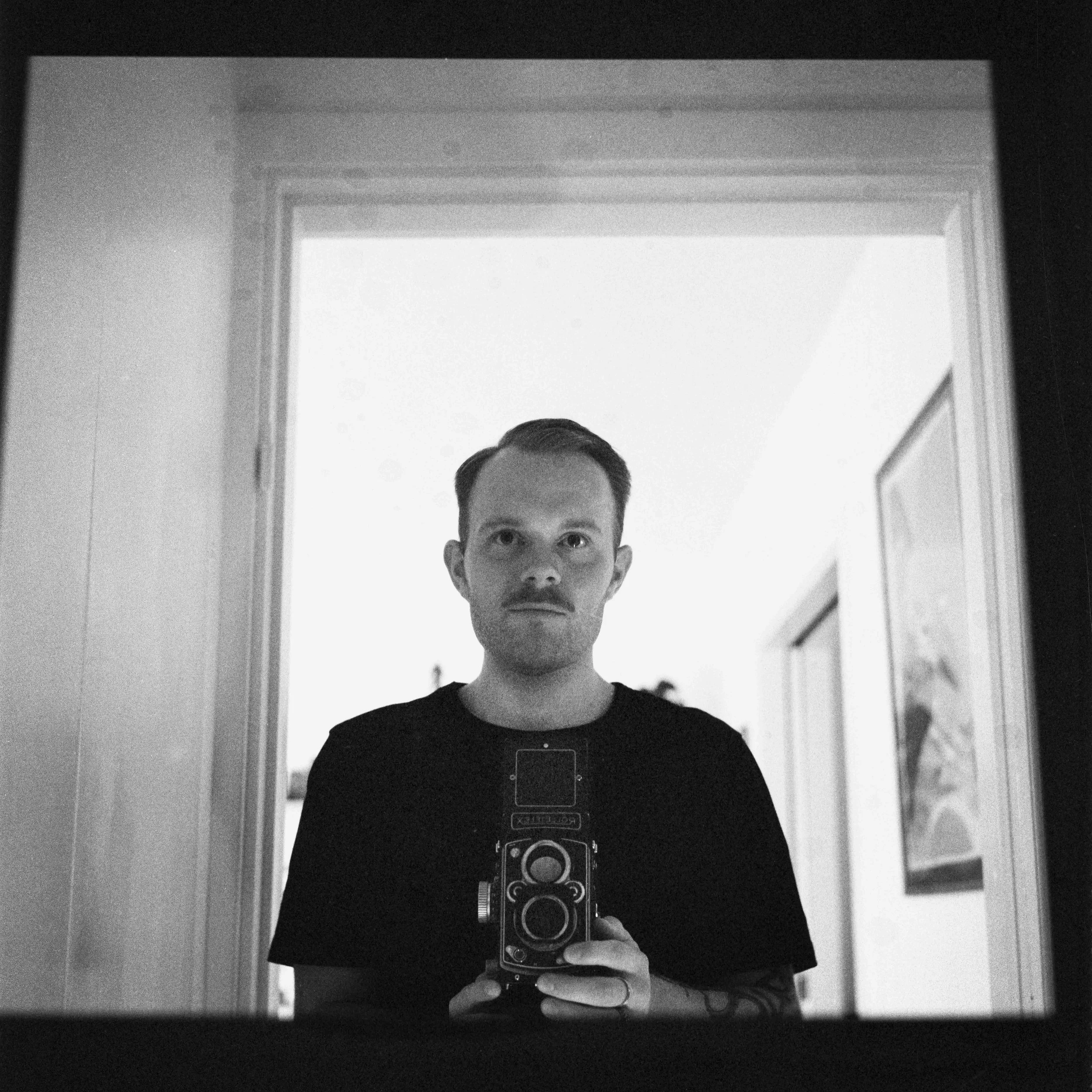 En man tar en svartvitt selfie med en gammal kamera i en spegel i ett rum.