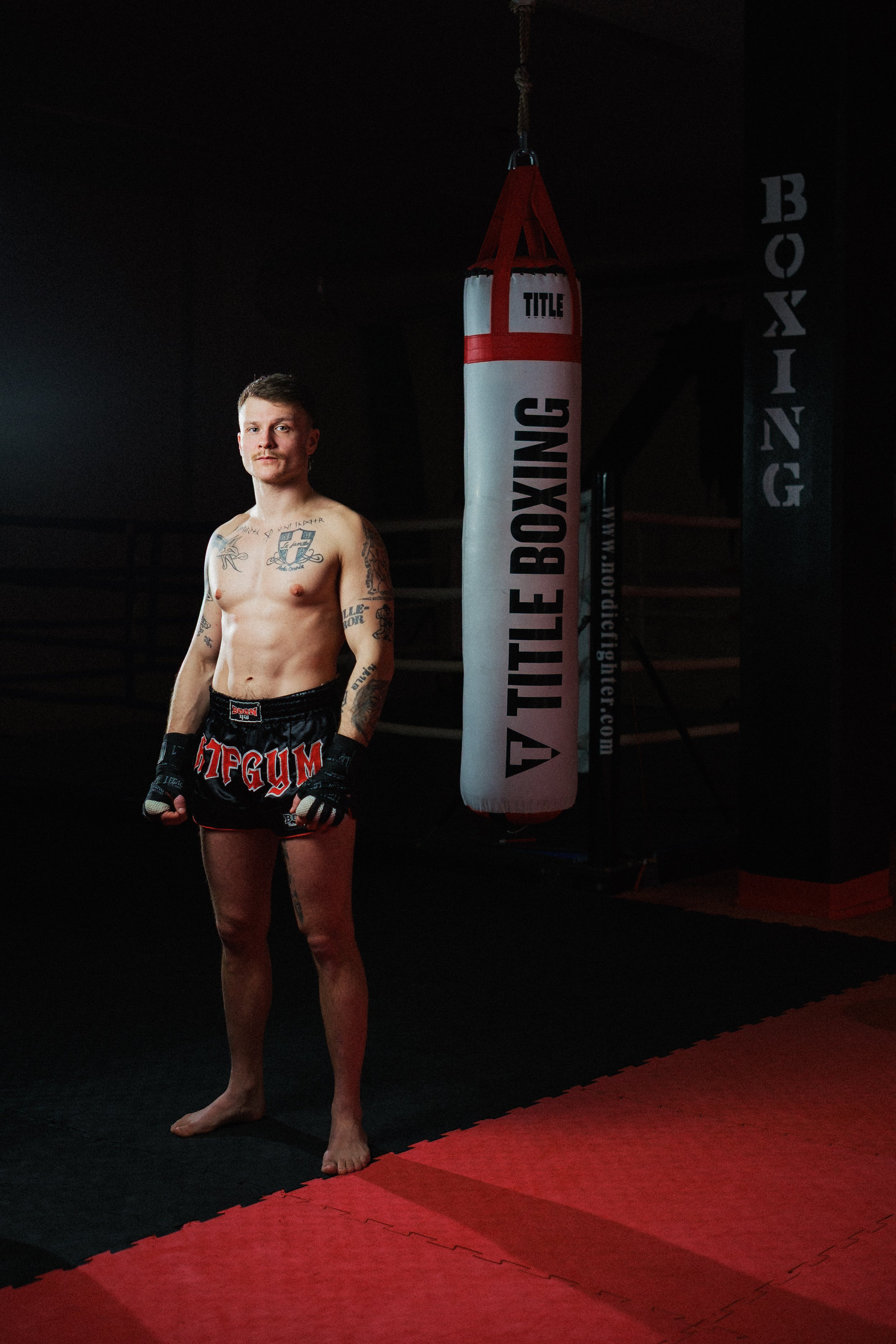 En muskulös man i kampsportkläder står i ett träningsrum bredvid en boxningssäck. Det är svart bakgrund och rummet har en matta på golvet.