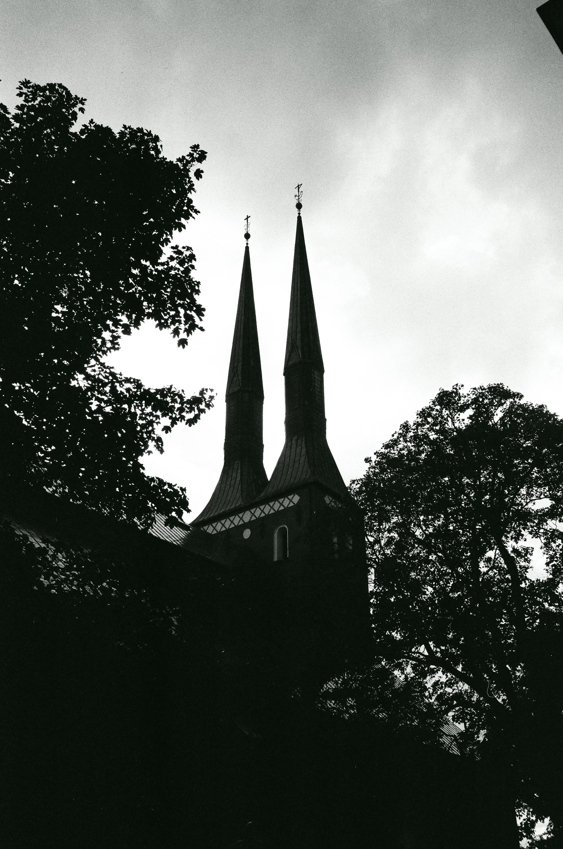 Svartvit bild av en kyrkas torn med två spira, omgiven av träd.