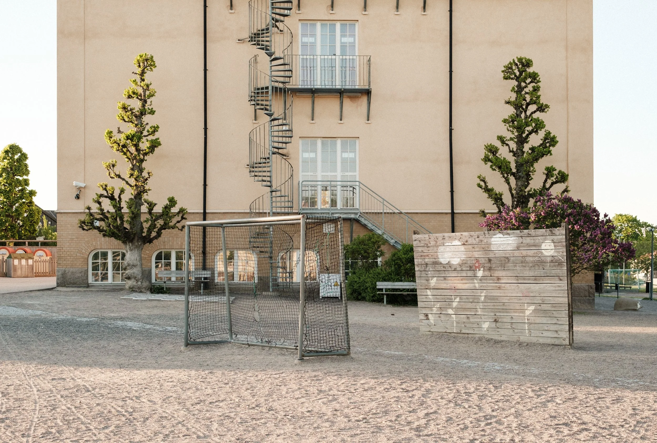 En tom lekplats med ett litet fotbollsmål, två träd, en vägg med en spiraltrappa, och buskar. Solen skiner, och det verkar vara en lugn dag.