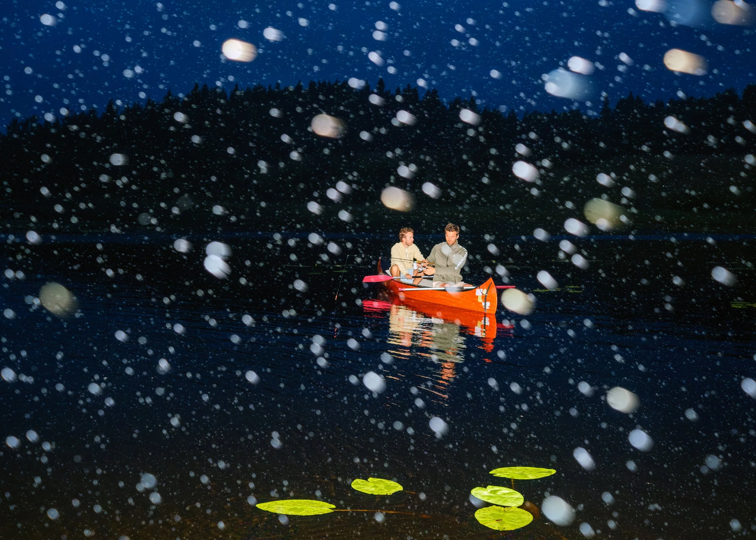 Två personer i en röd båt på en mörk sjö omgiven av en skog, snö faller och reflekteras i vattnet under en mörk himmel.