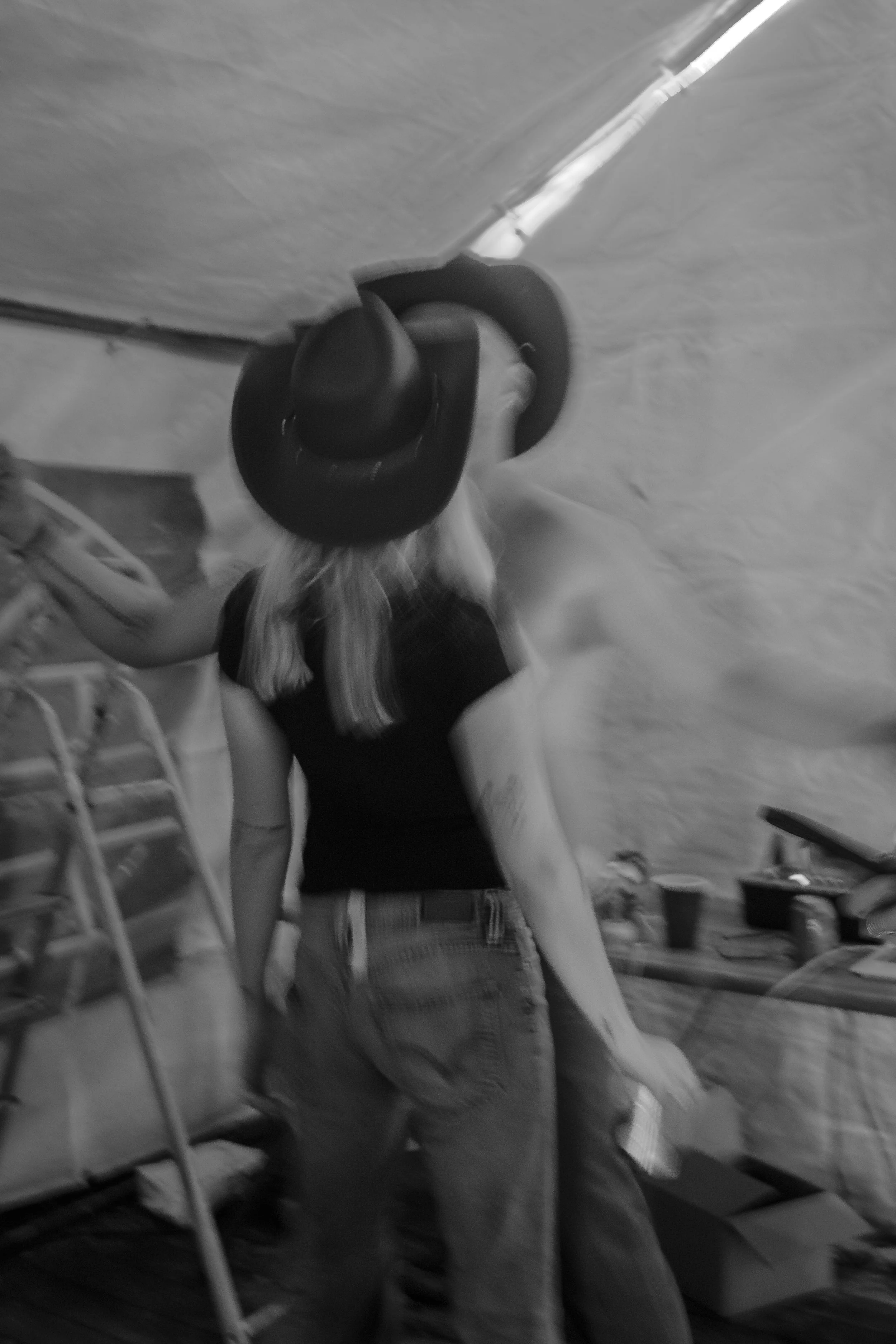 En kvinna som bär en svart hatt och svart T-shirt, sett bakifrån, i ett rum med snedtak och olika föremål, inklusive en stege och bord.