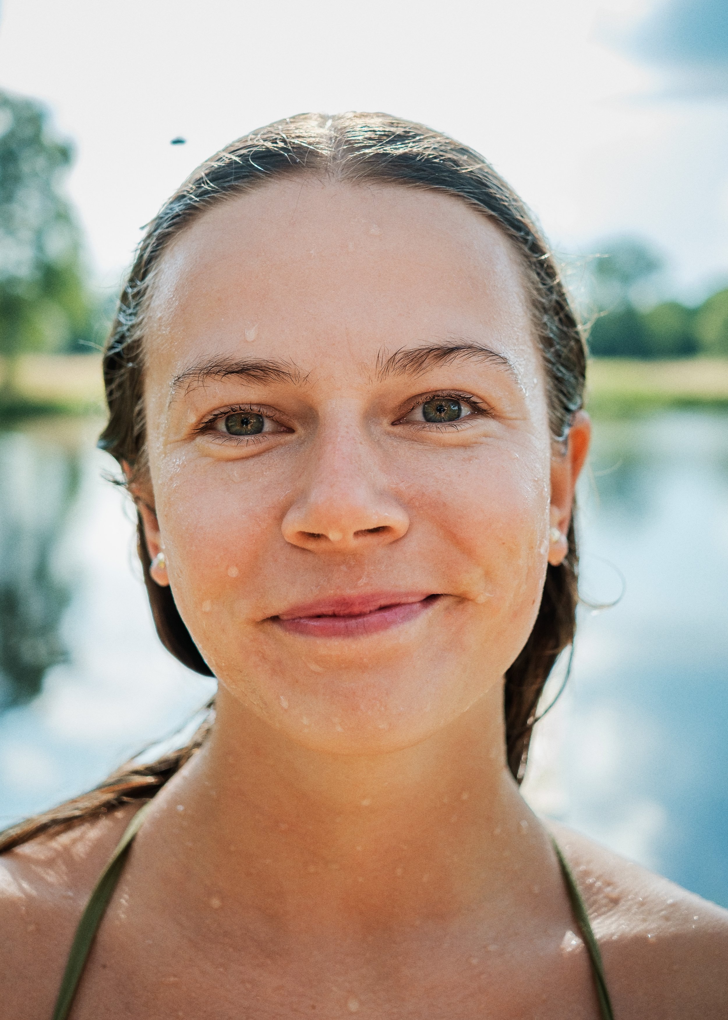 En ung kvinna med blött hår, ler och står utomhus nära en vattenspegel.