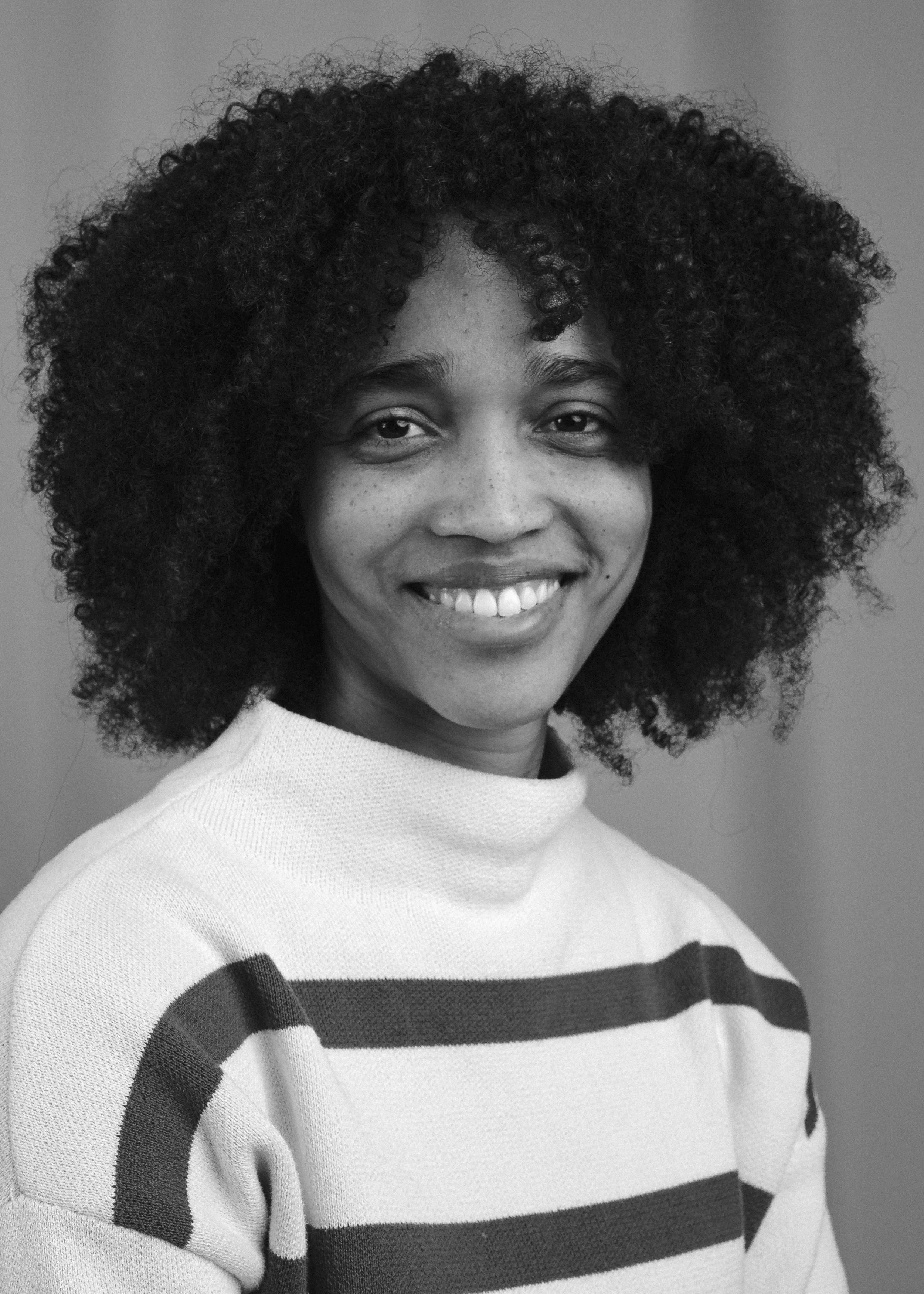 Svartvitt porträtt av en kvinna med lockigt hår, leende, iklädd en ljus tröja med breda mörka ränder, mot en enkel bakgrund.