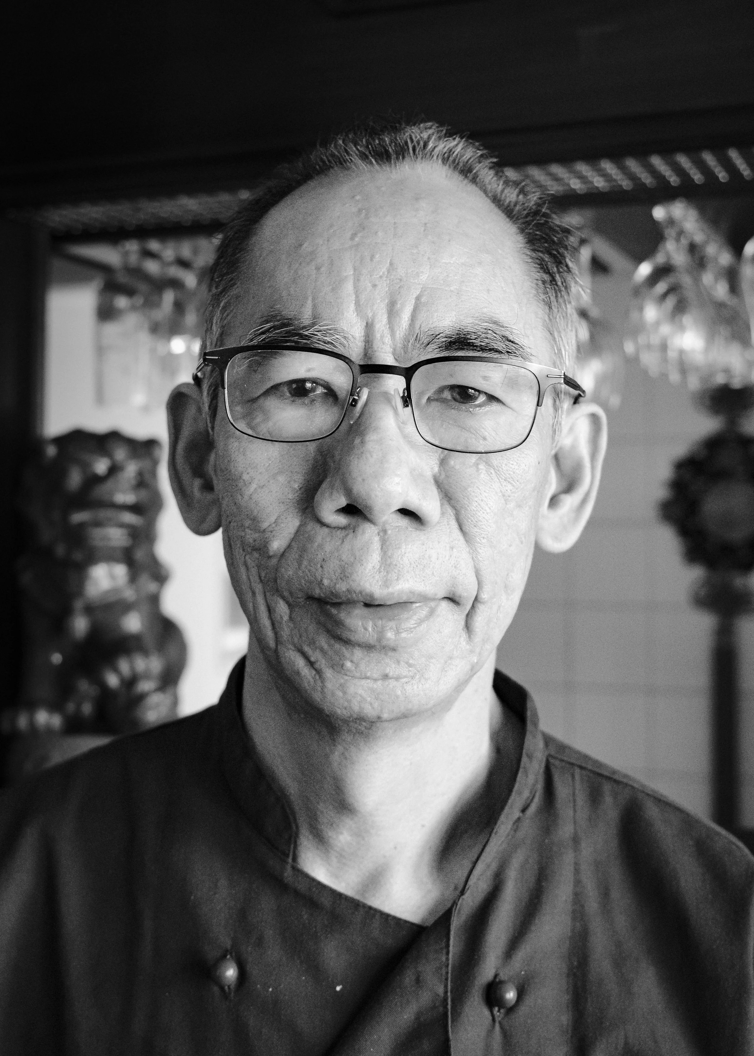 Svartvit bild av en man med glasögon, kort hår och ett leende, troligen i en restaurang eller ett kök, med dekorativa föremål i bakgrunden.