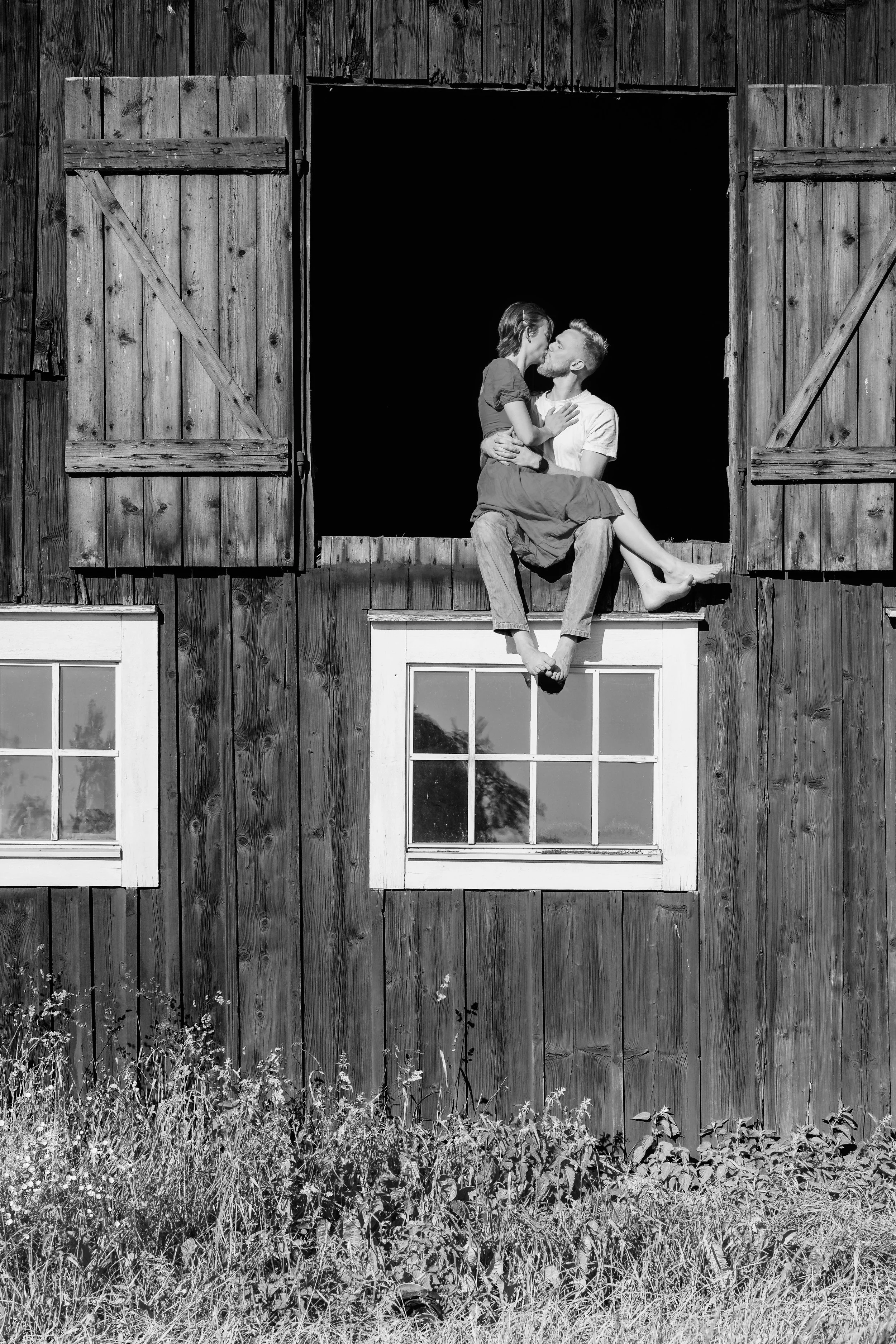 Ett svartvitt foto av ett par som pussas på ett fönsterbräde av en trävilla, med stängda fönster och öppet fönster utan glas.