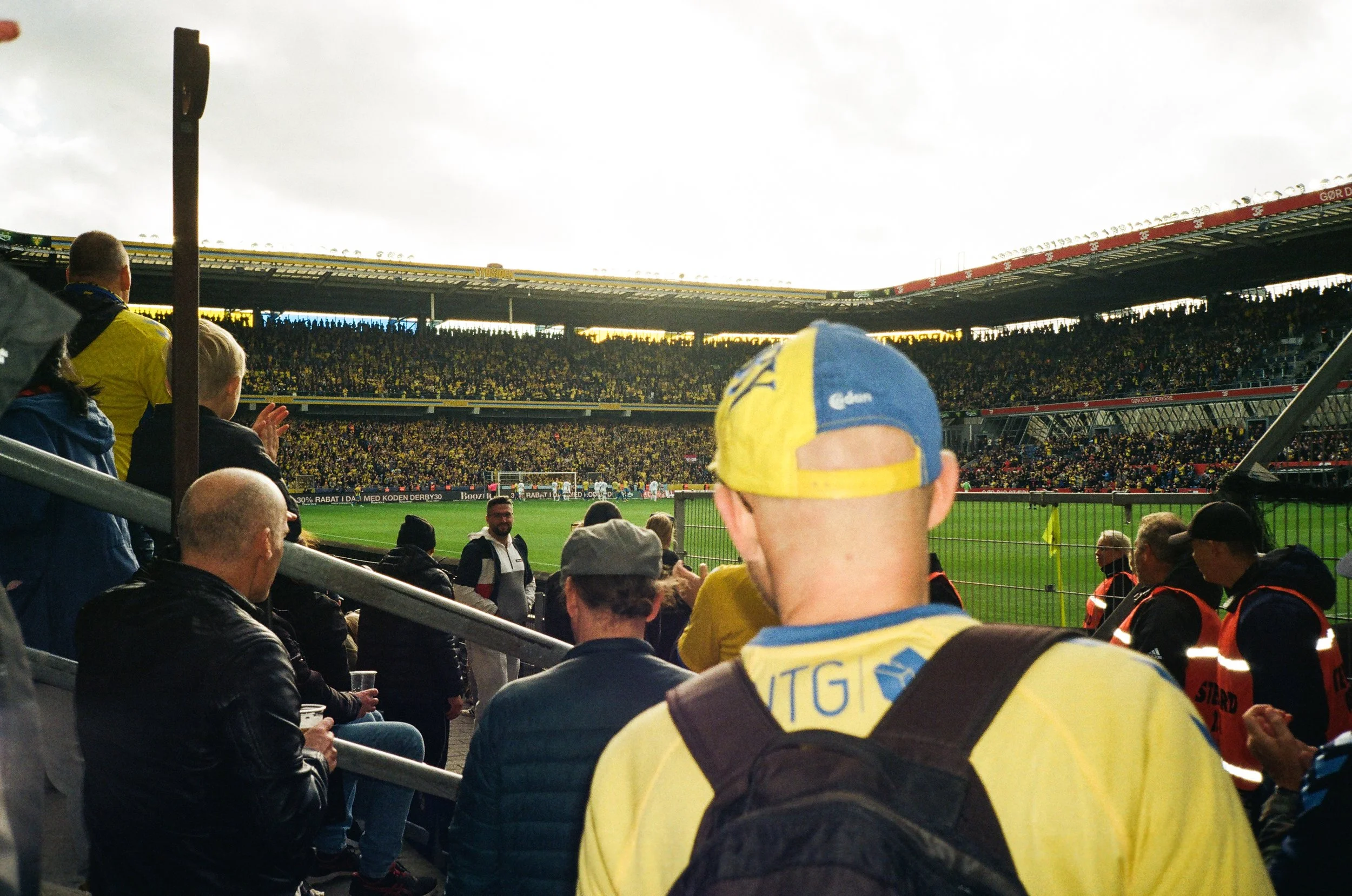 Fodboldstadion med fans som tittar på en match, många är klädda i gult och blått, inklusive en person i förgrunden med en gul och blå keps och tröja.