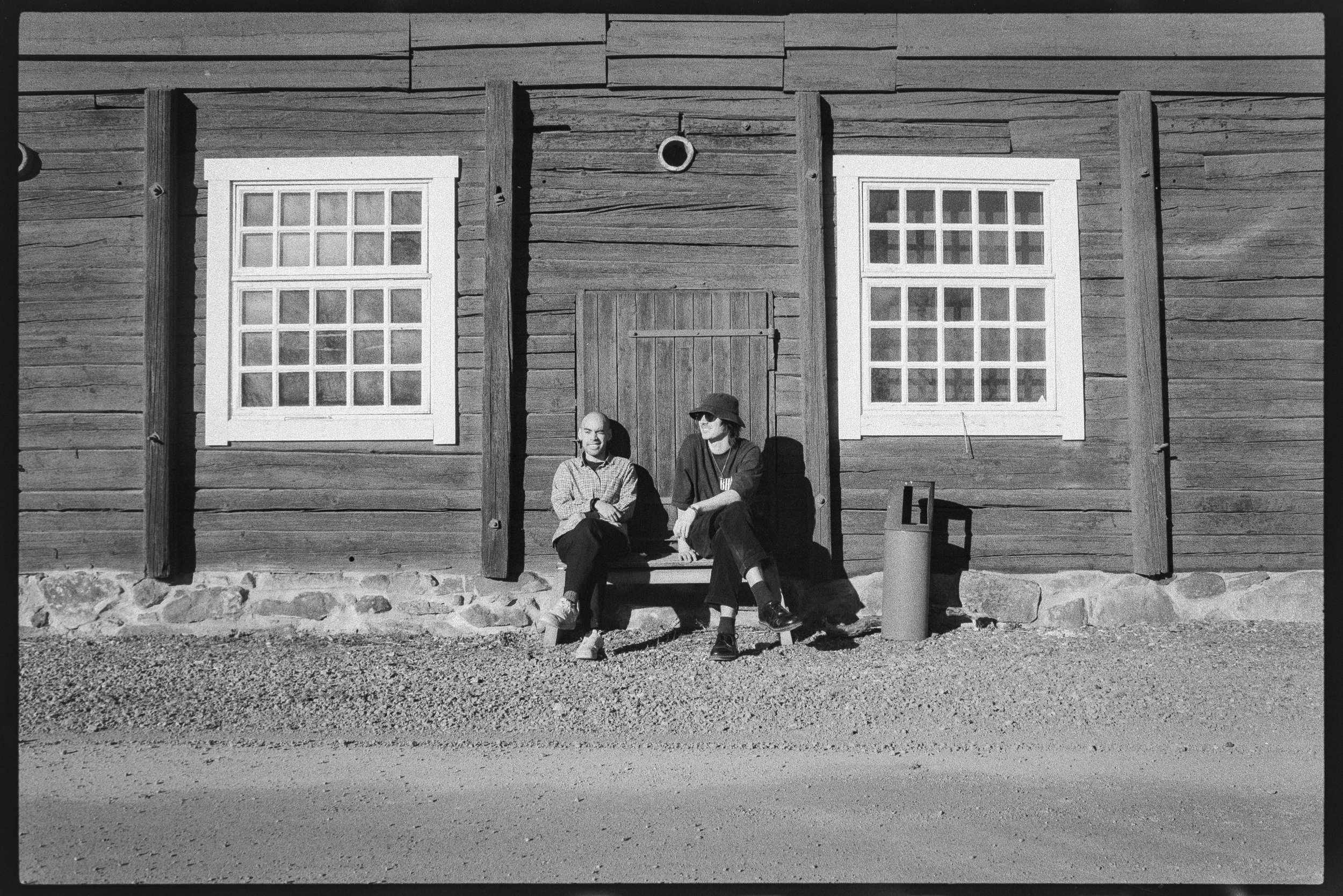 Två personer sitter på en bänksläd detaljerade, framför en trähusvägg med två stora fönster och en liten dörr i mitten. Fönstren har vita ramar och många små rutor. Det finns en liten soptunna till höger om personerna.