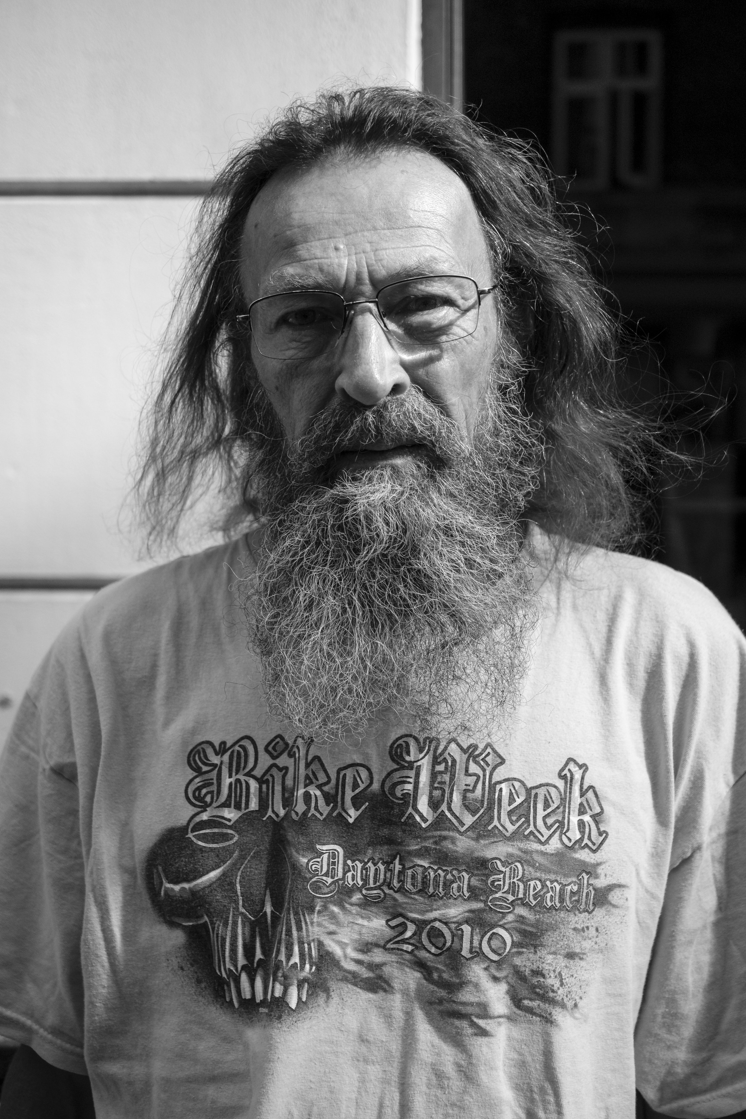 En man med glasögon, ett långt grått skägg och hår, som bär en T-shirt med texten 'Bike Week Daytona Beach 2010' och en skalle. Bilden är i svartvitt.