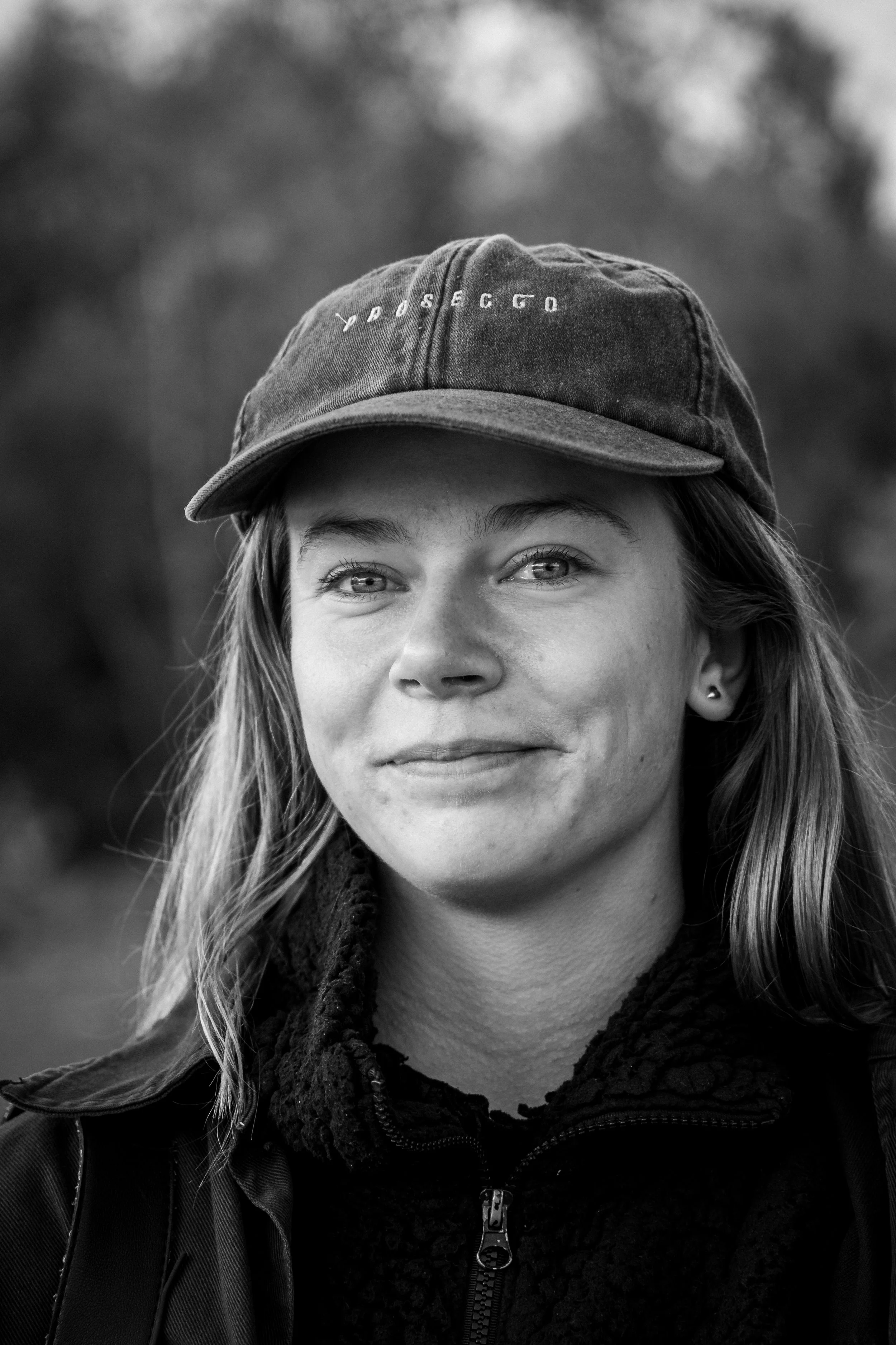 Svartvitt foto av en leende kvinna med keps och jacka utomhus.