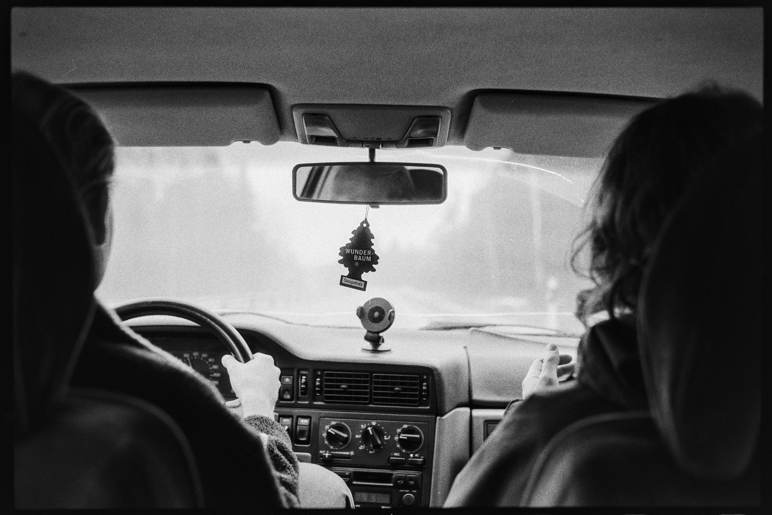 Svartvit bild av två personer i en bil, sedd från baksätet. Man ser ratten, instrumentbrädan och främre sidorutan. En luftfreshener i form av ett granträd hänger i backspegeln med texten 'WUNDERBAUM'.