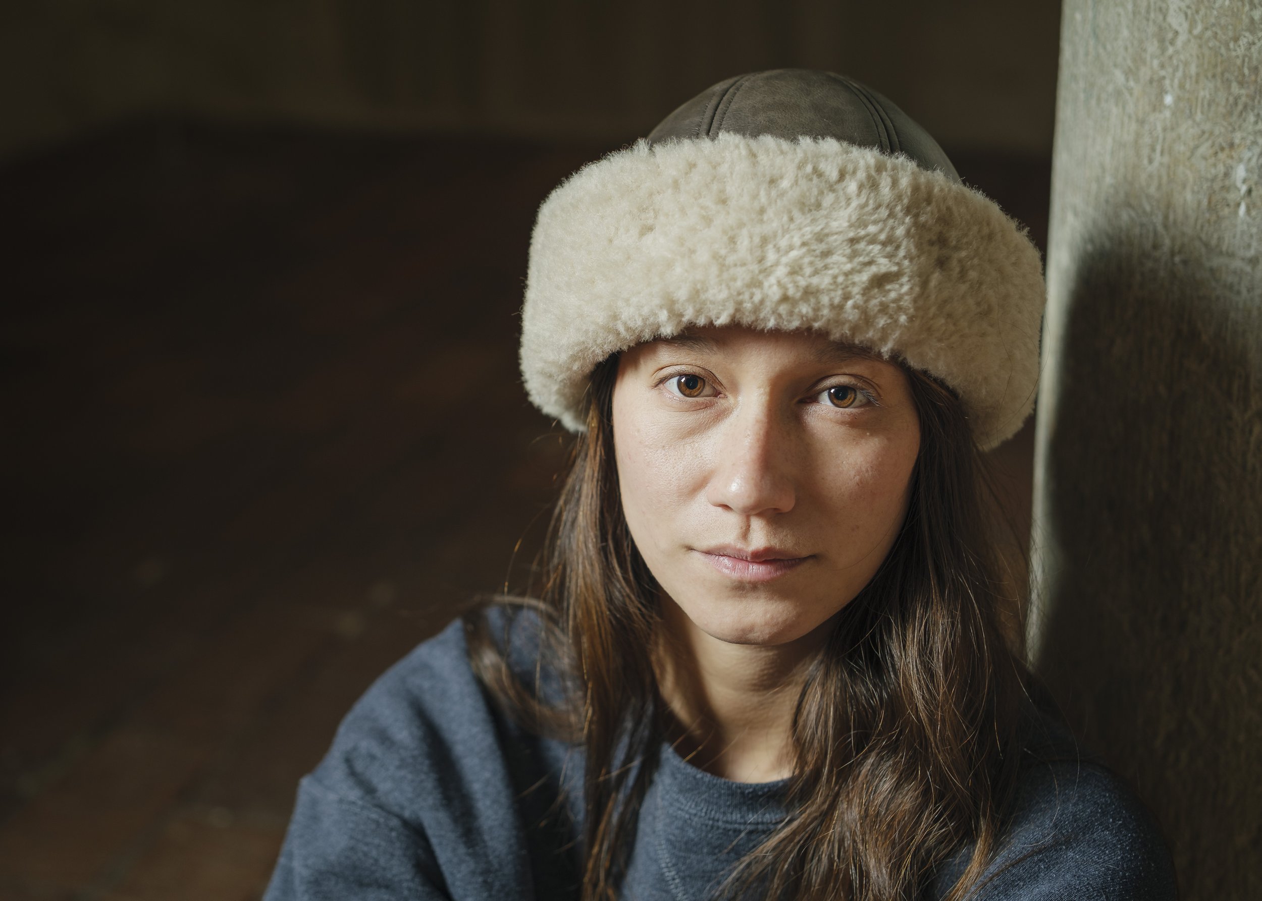En ung kvinna med långt brunt hår, som bär en beige, ullig mössa med en dold luvhatt, sitter intill en vägg av trä, tittar mot kameran.