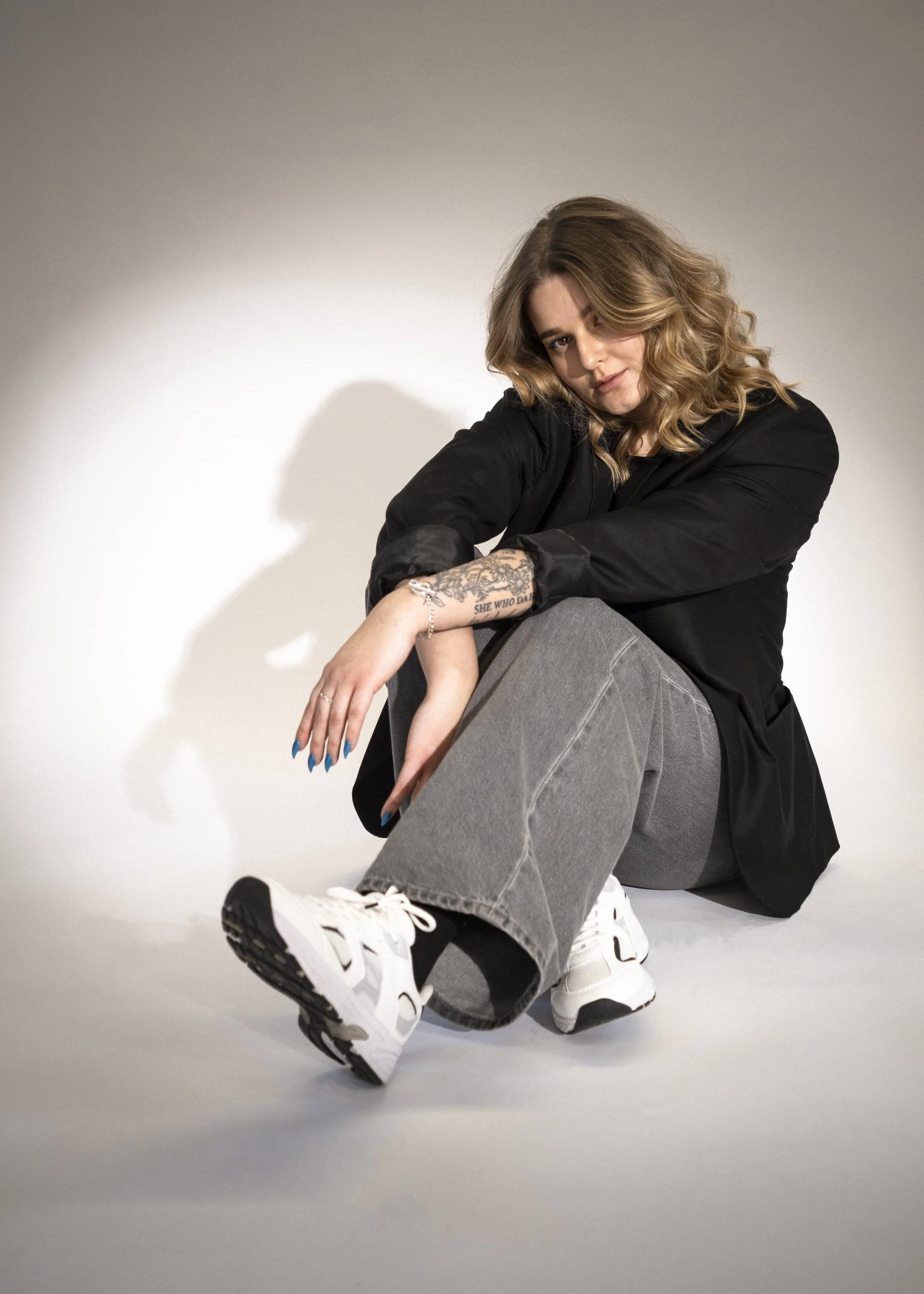 En ung kvinna med lockigt hår, svart jacka, gråa jeans, och vita sneakers, som sitter på golvet och tittar in i kameran.