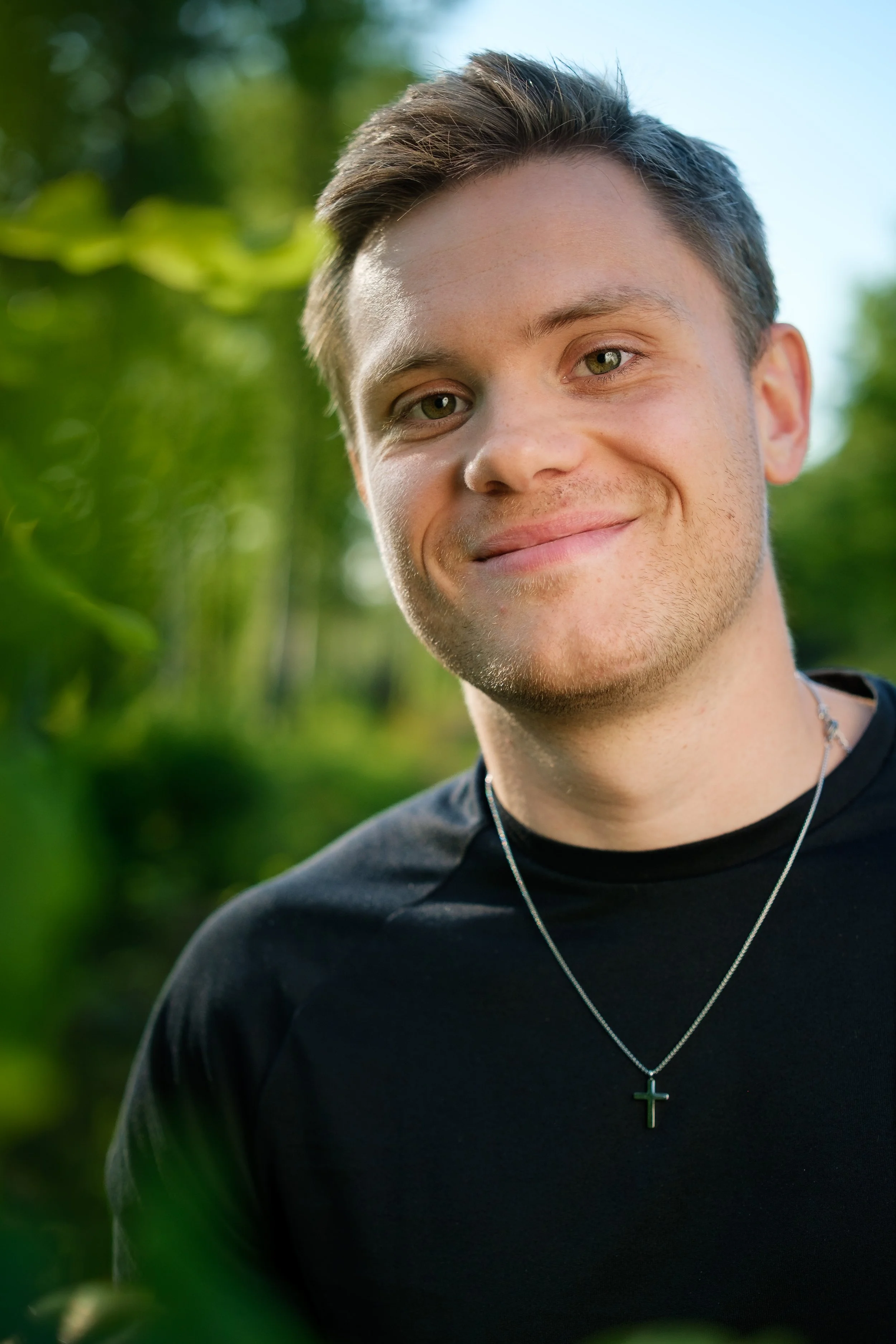 En ung man med kort, mörk hår och en rundad skäggstubb ler i en grön, utomhusmiljö.