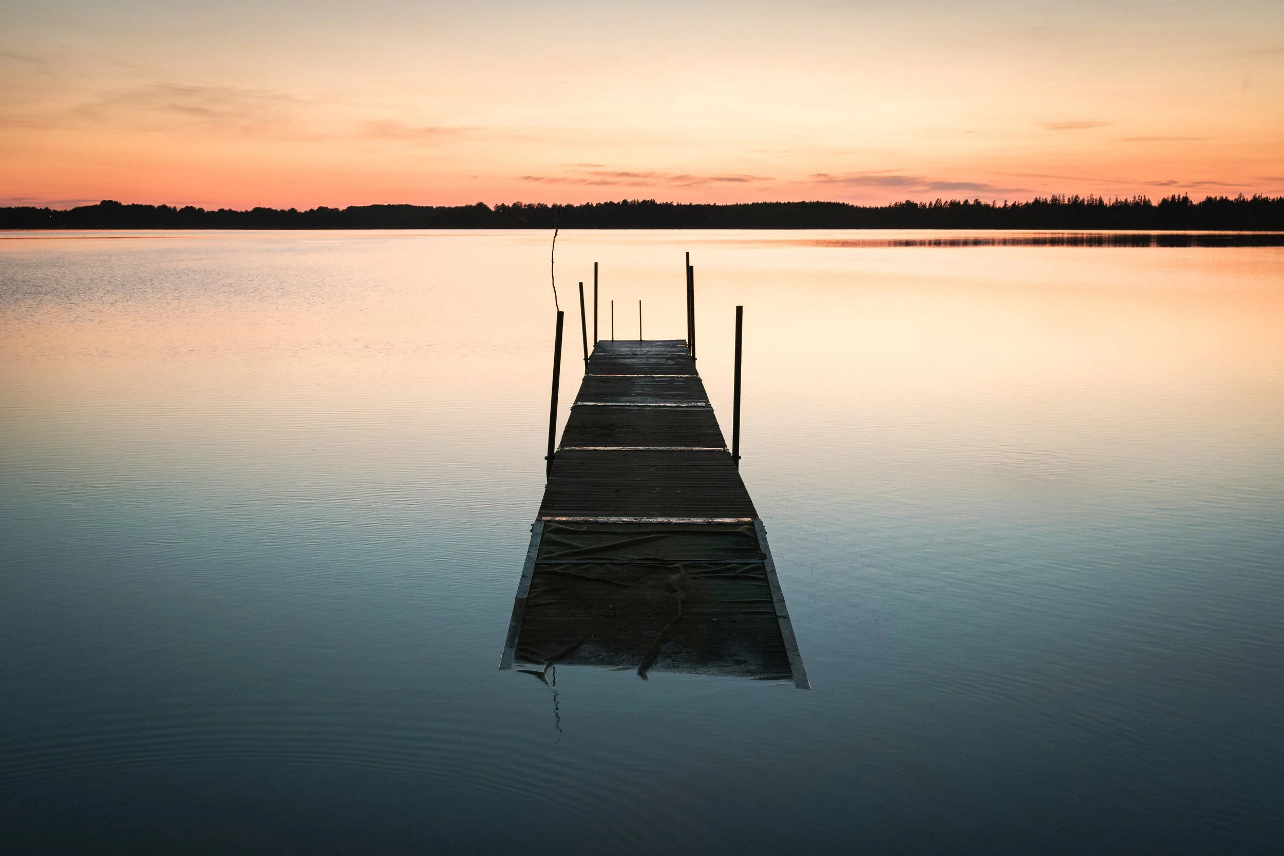 En träbrygga som sträcker sig ut i en stilla sjö under en solnedgång, med en rosa och orange himmel och en mörk siluett av träd i bakgrunden.