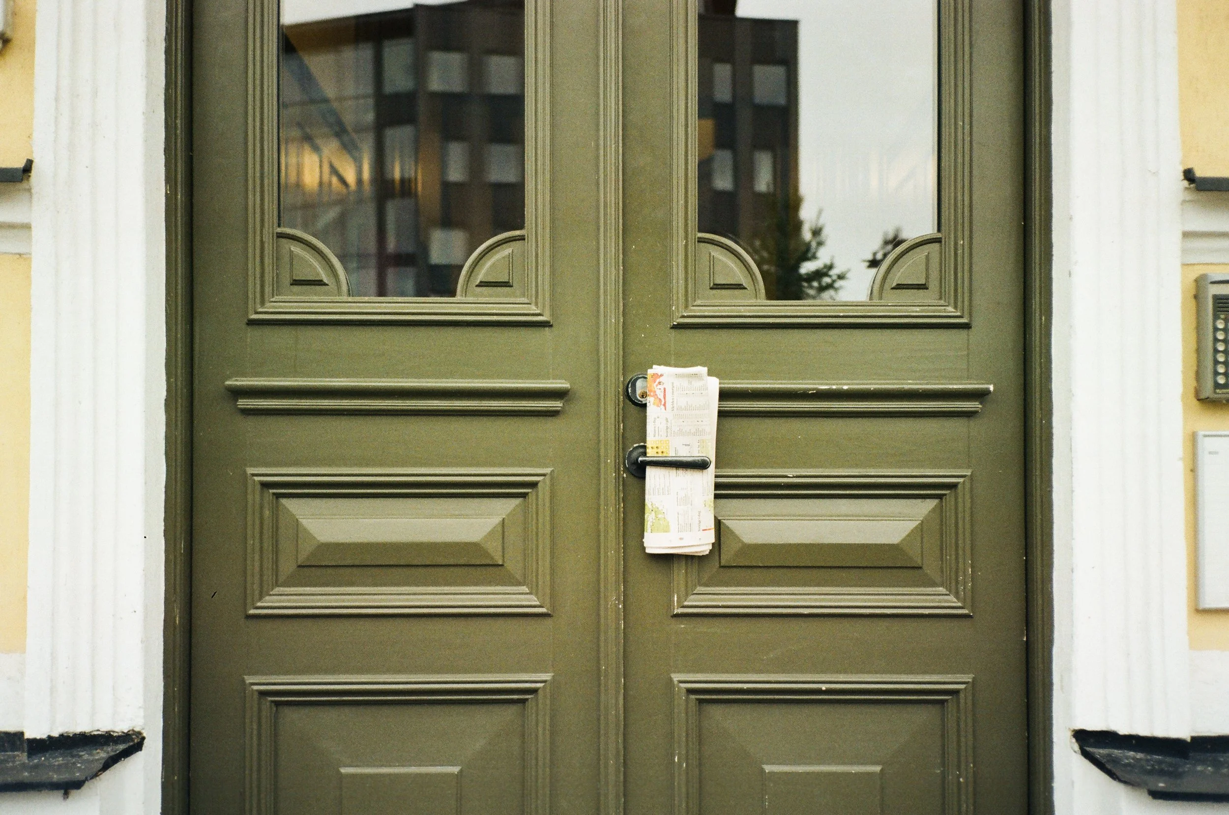 Stängda gröna dubbeldörrar med glasfönster och klistremerke, omgivna av en gul vägg.